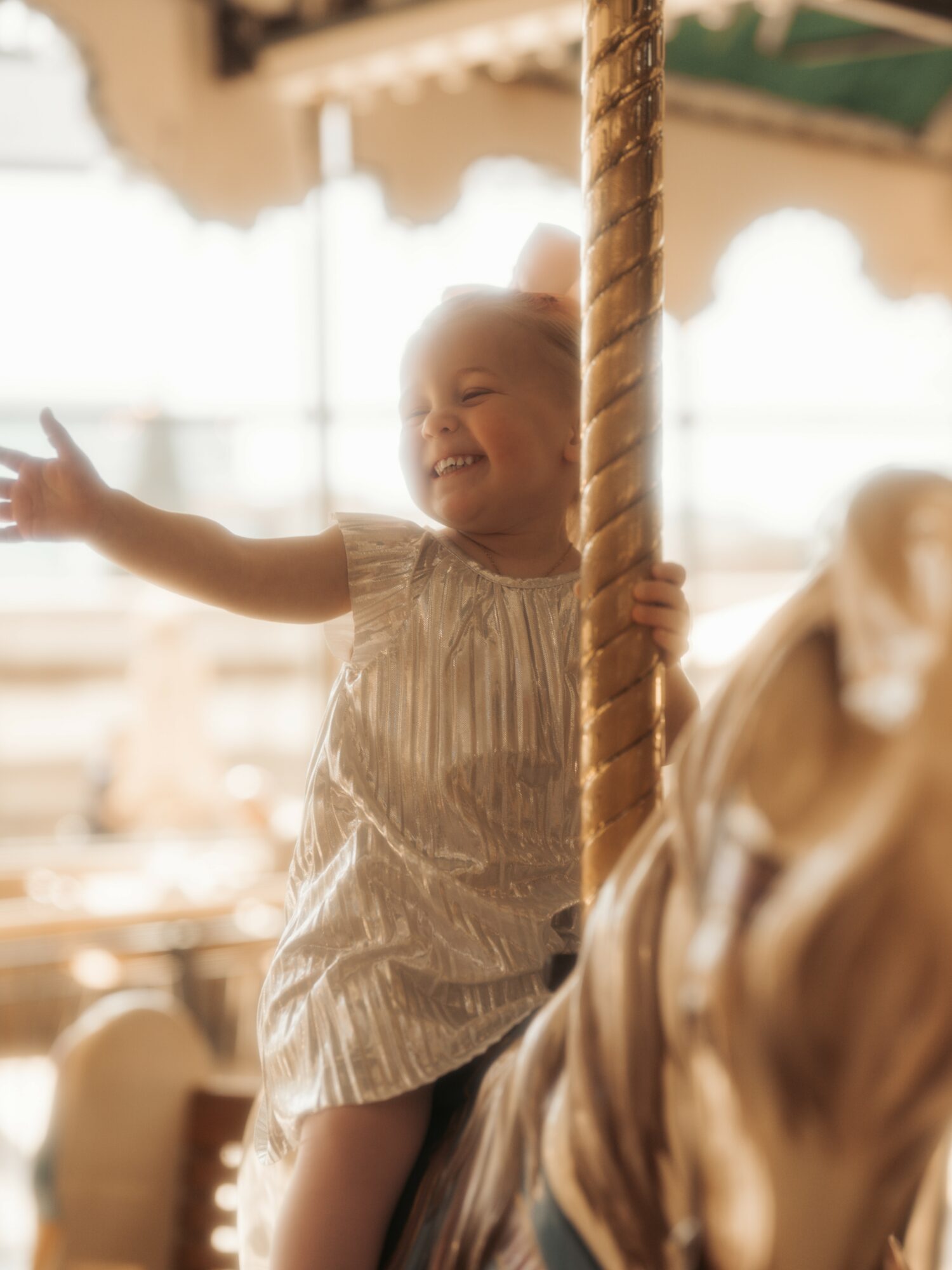 Child smiling and reaching out while holding a rope on a carousel, with blurred background and another person nearby.