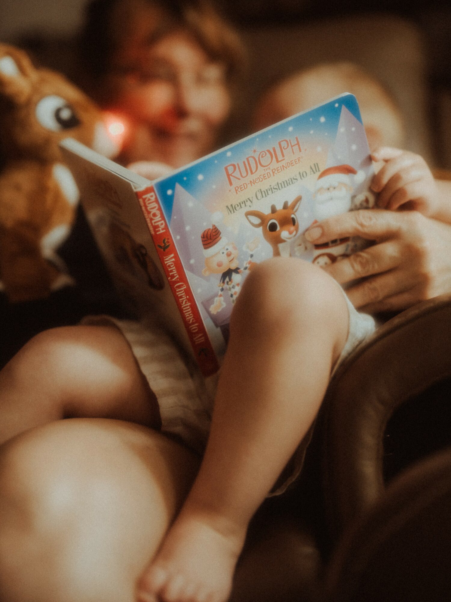 Child holding a Rudolph-themed book, with a reindeer plush toy nearby, smiling in a cozy setting.