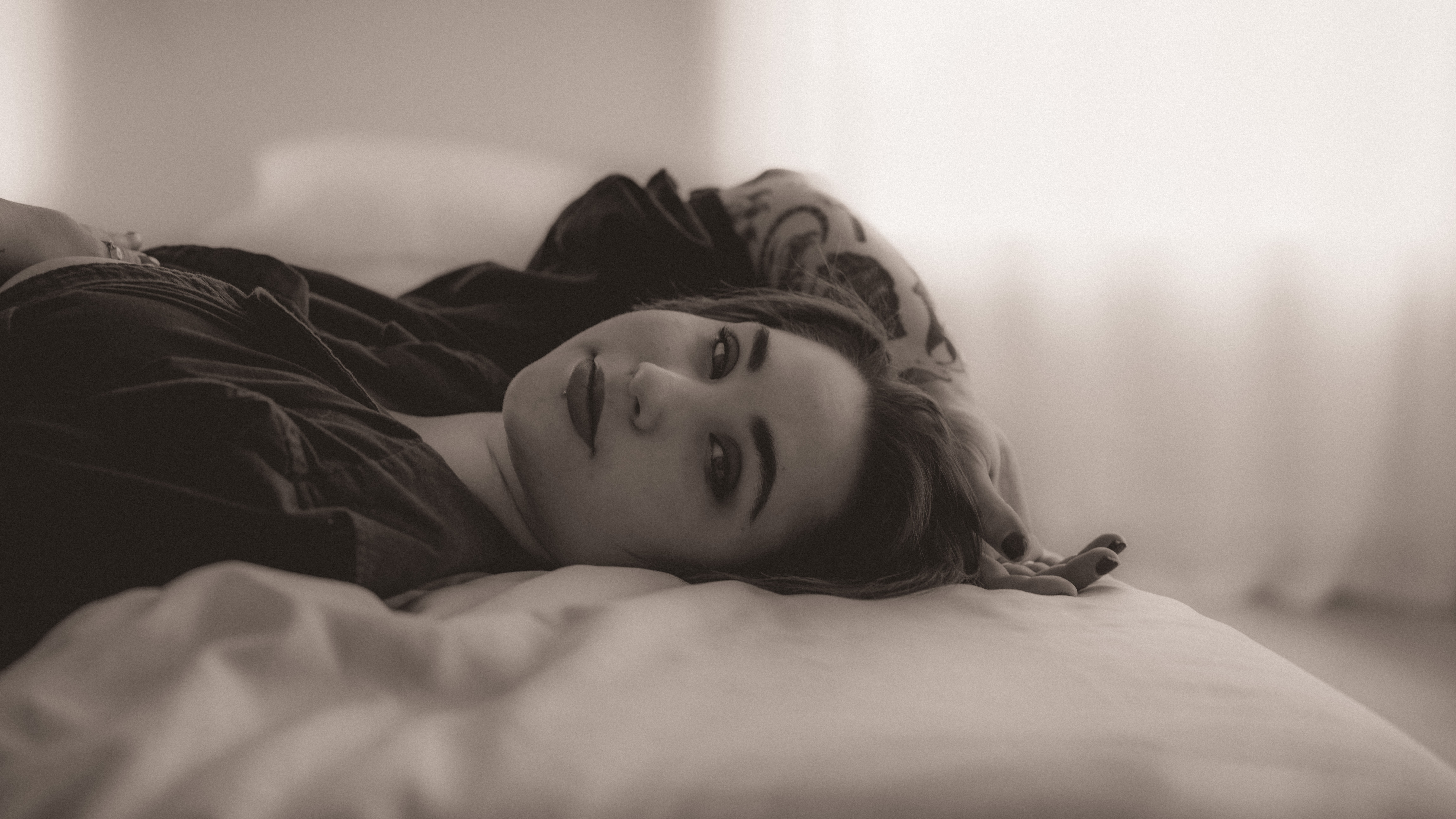 Woman lying on bed with head resting on pillow, looking at camera, in a softly lit room, black and white photo.