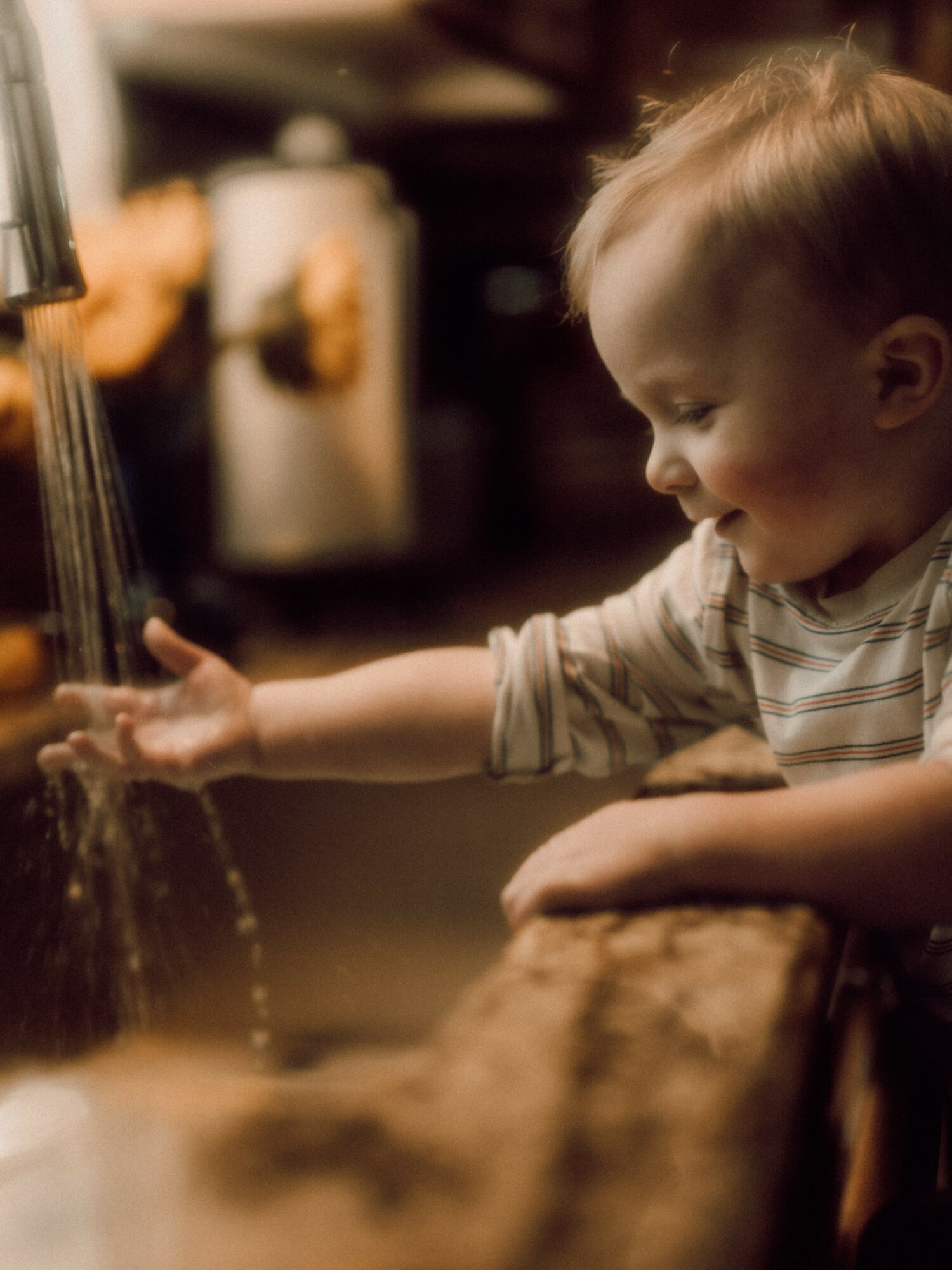 Child reaches for water at a sink, smiling, with a blurred background of kitchen items.