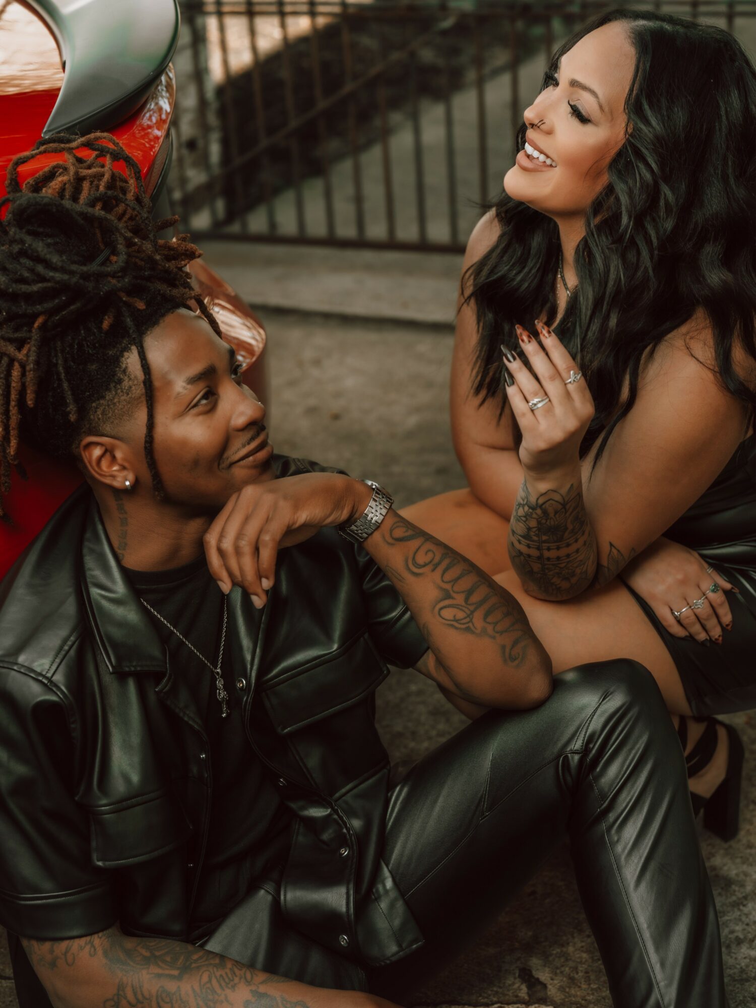 Two women smiling and talking outdoors, one with dreadlocks and the other with long dark hair, sitting on the ground.