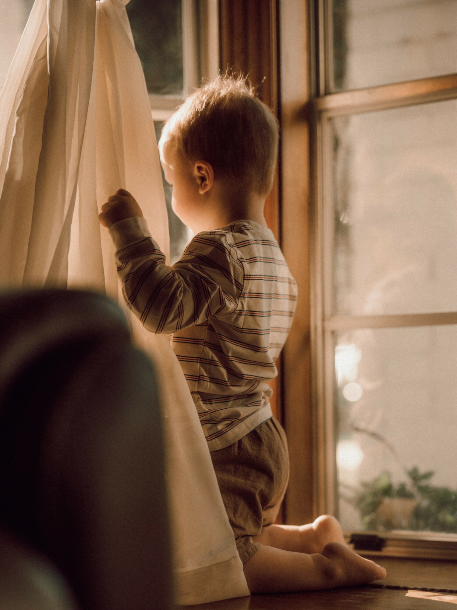 Child with short hair looking out a window, sitting on a windowsill, with sunlight illuminating the scene.