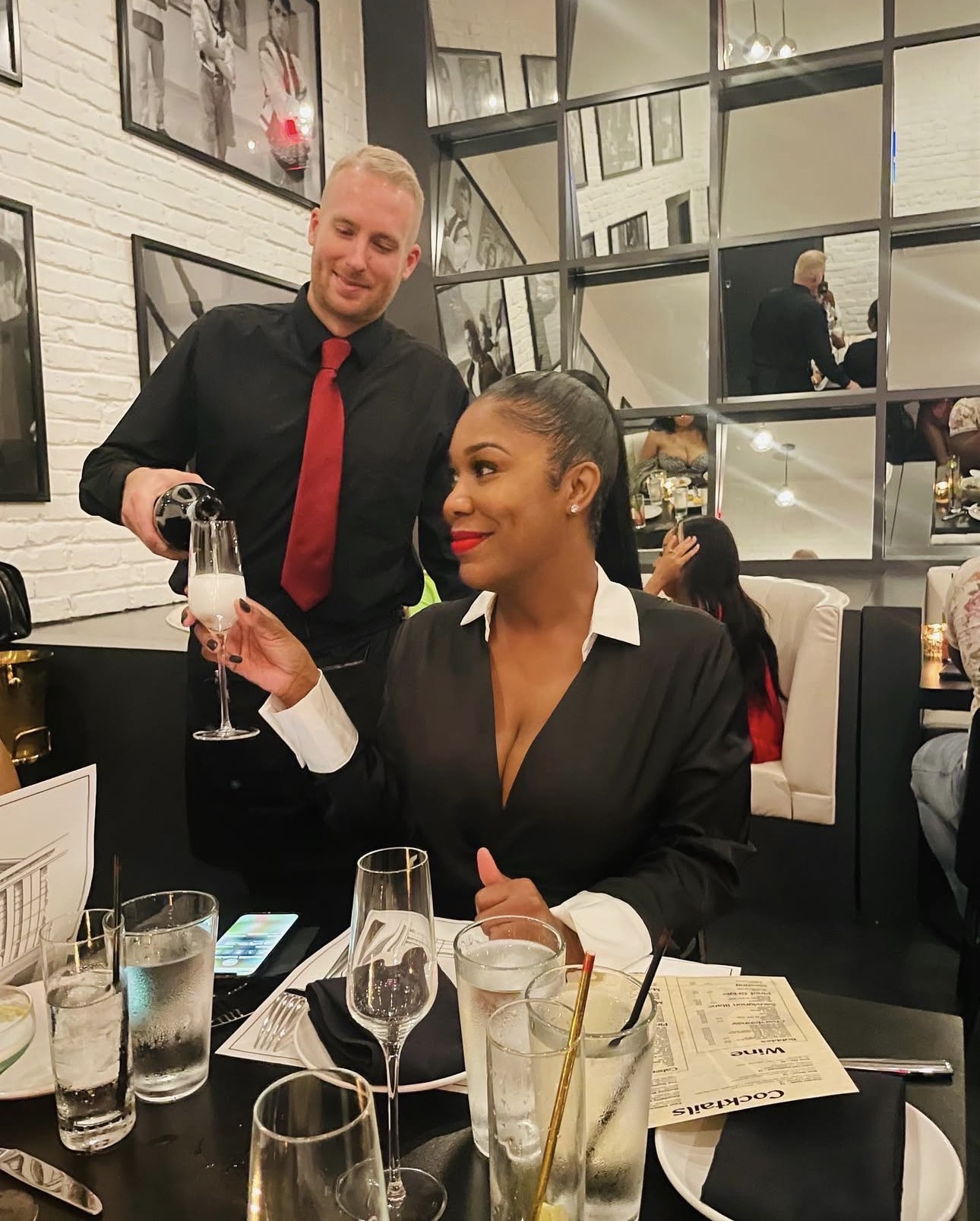 A woman in a black blouse with a white collar holds a glass of champagne, seated at a restaurant table with a man standing behind her. The woman has dark hair and red lipstick, and the man has short blond hair, a beard, and is wearing a black shirt with a red tie. The table has drinks, a menu, and a smartphone. The background shows a modern restaurant interior with mirrors and framed pictures.