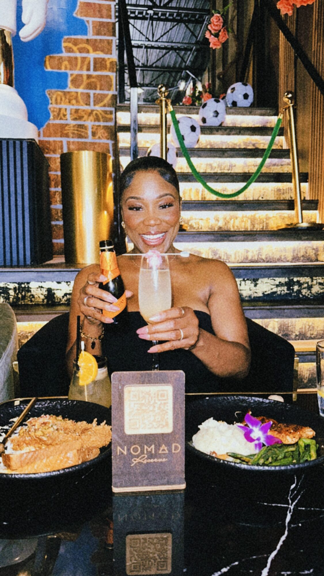 Smiling woman holding two drinks at a bar with snacks, stairs, and decorations in the background.