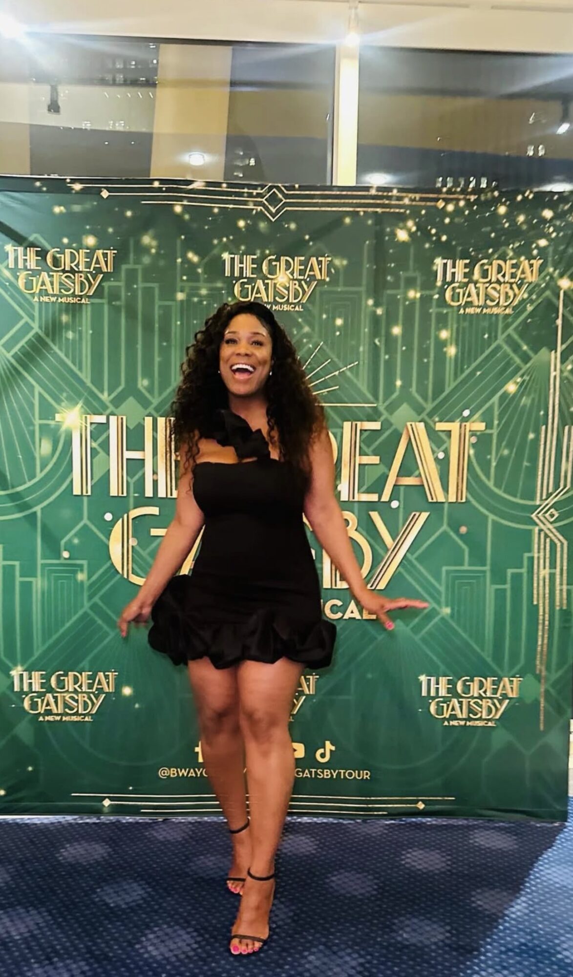 Woman with curly hair in a black dress standing in front of a green backdrop with gold text and patterns.
