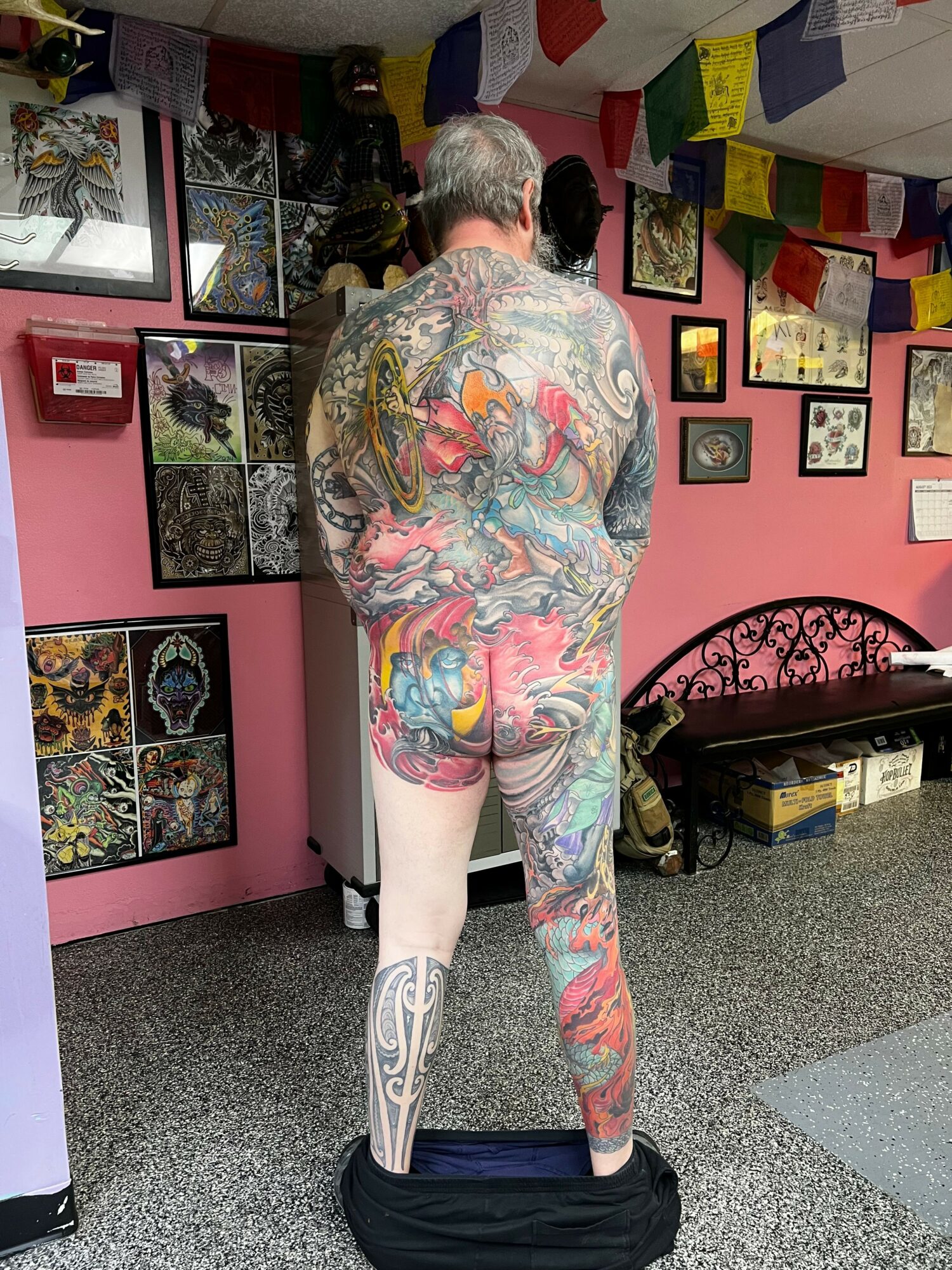 Person with colorful tattoos standing in a room with pink walls and framed pictures, facing away from the camera.