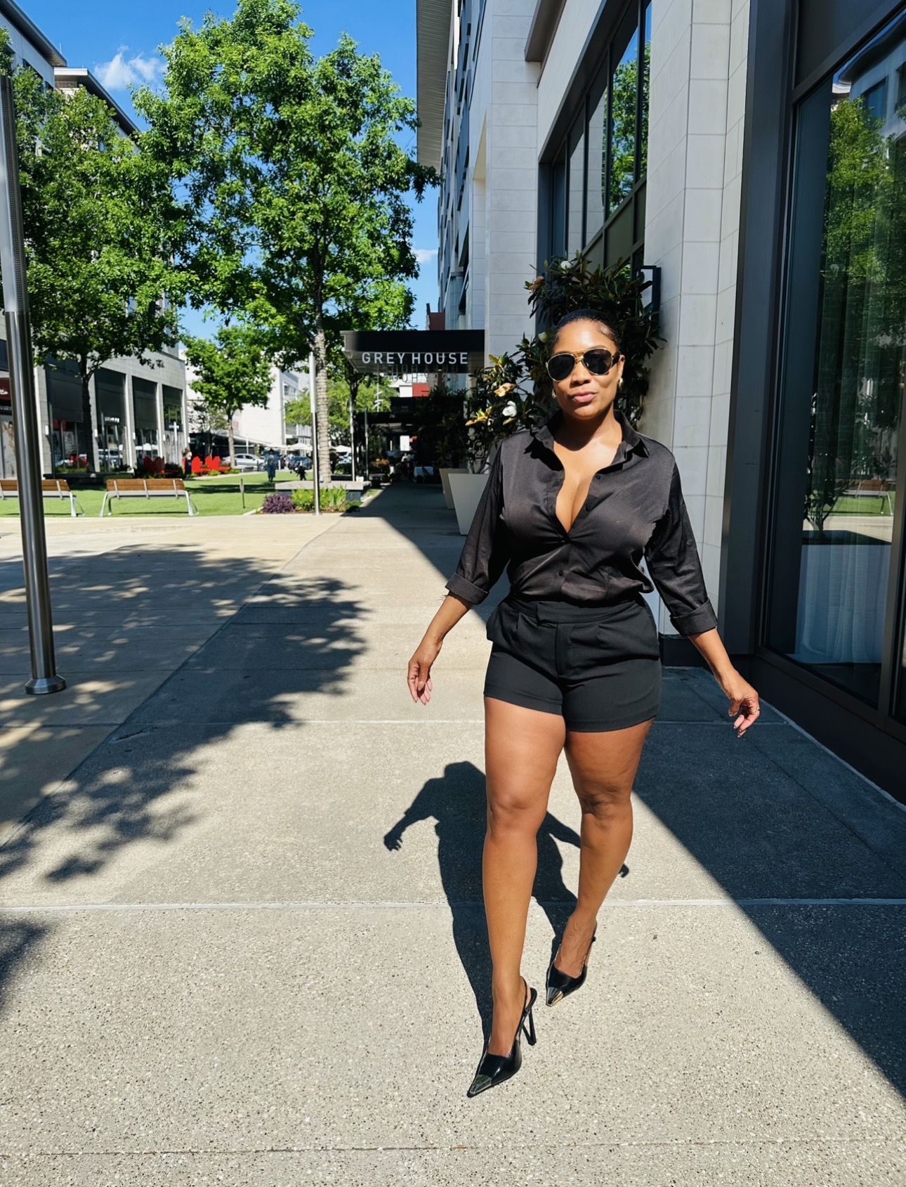 Woman in black outfit walking on sidewalk with trees and buildings in background.