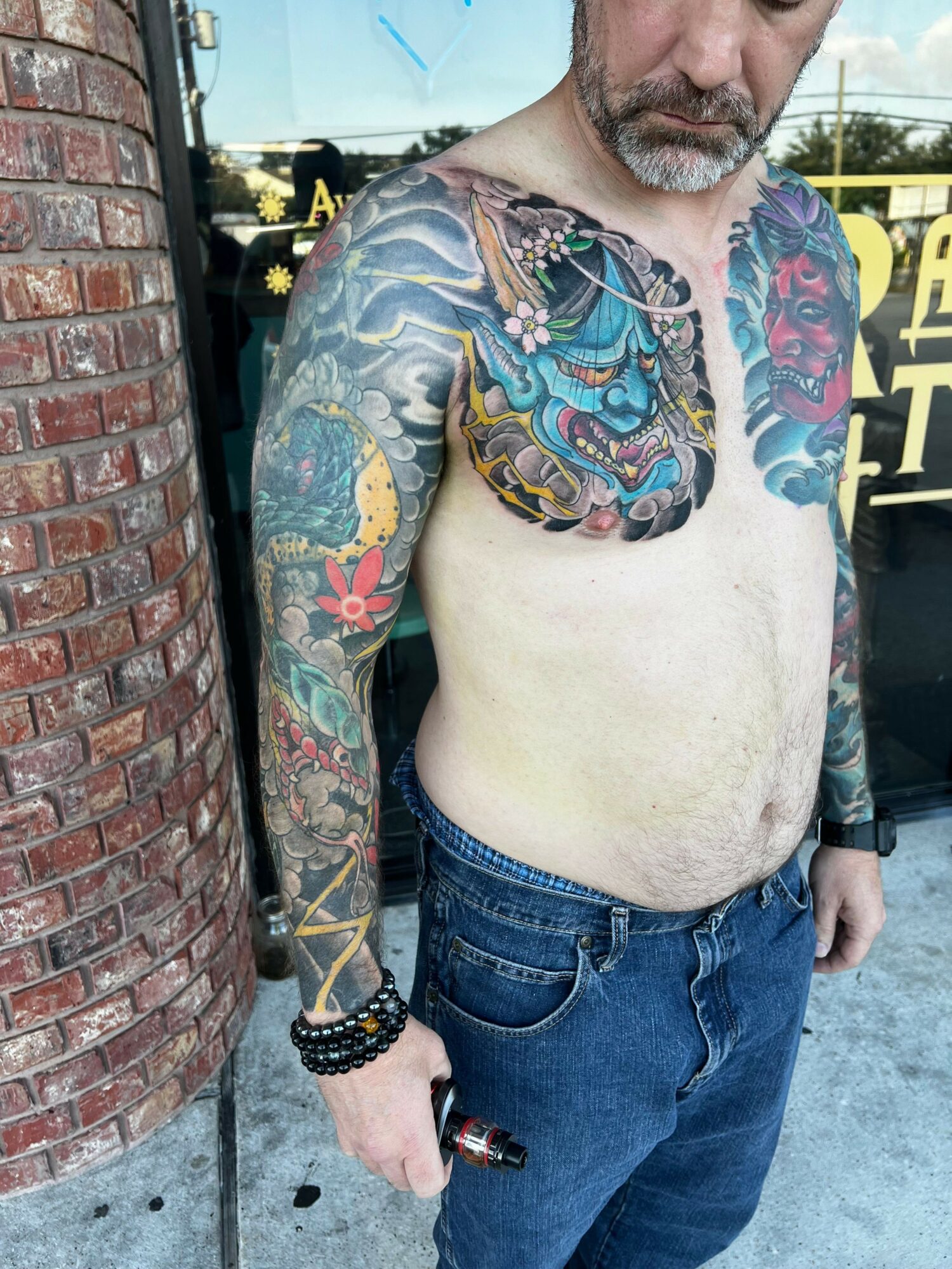 Man with colorful tattoos on arms and chest, wearing jeans, standing outdoors near a brick wall.