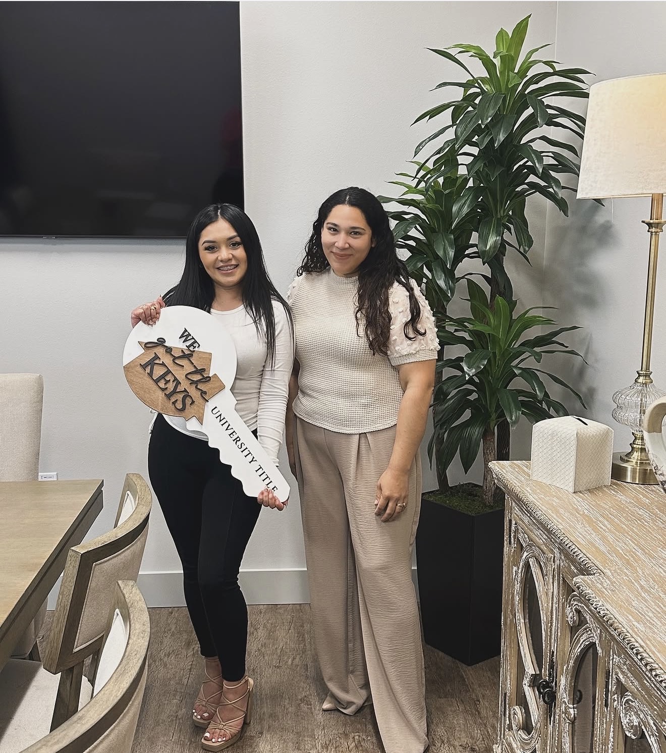 Two women standing indoors, smiling, with a large plant and a lamp behind them, in a well-lit room.