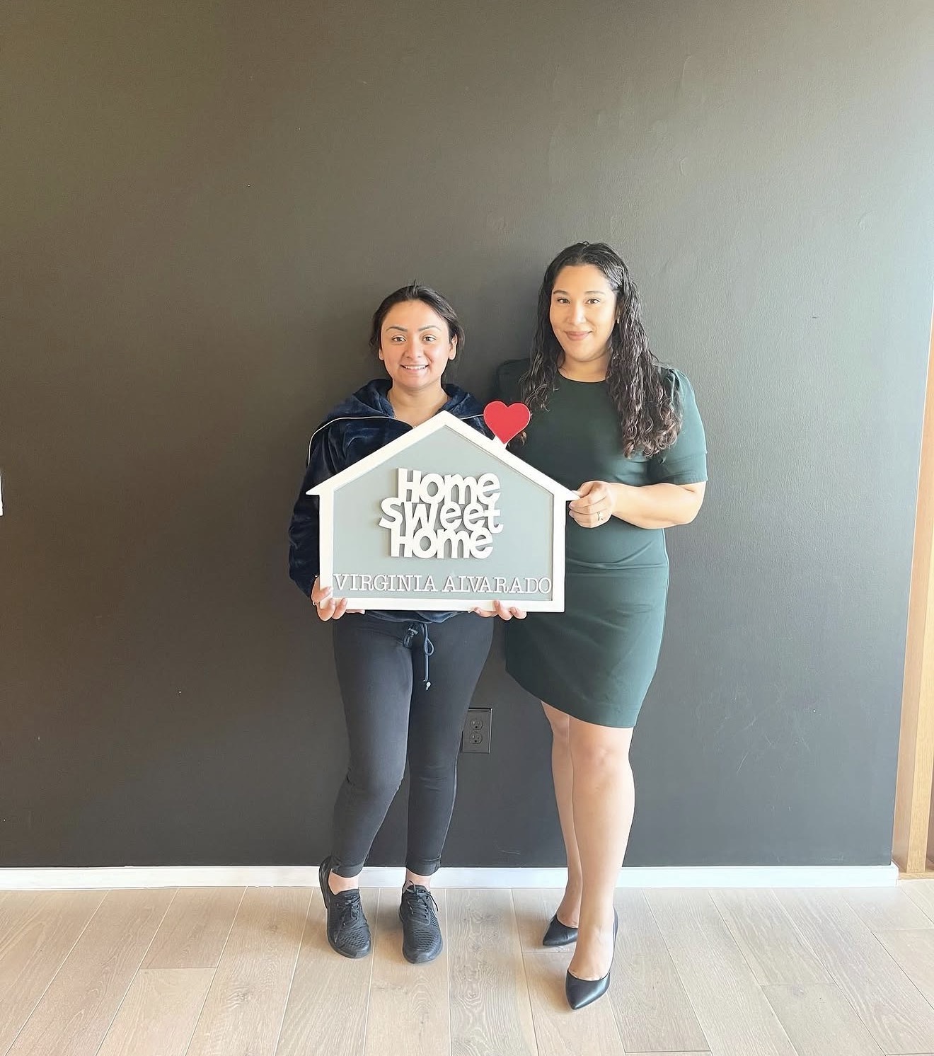Two women standing against a dark wall, holding a sign that says