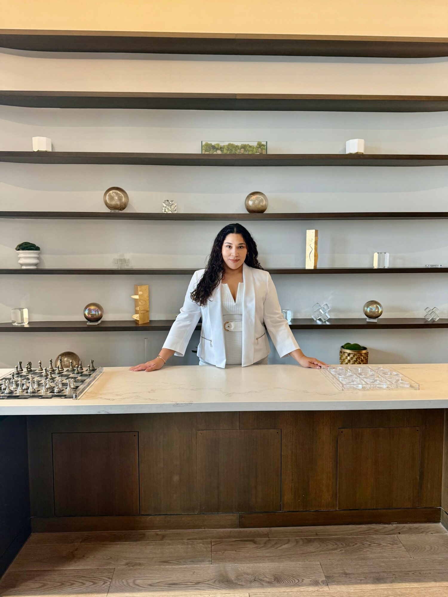 Woman standing behind a kitchen counter with shelves and decorative items in the background.