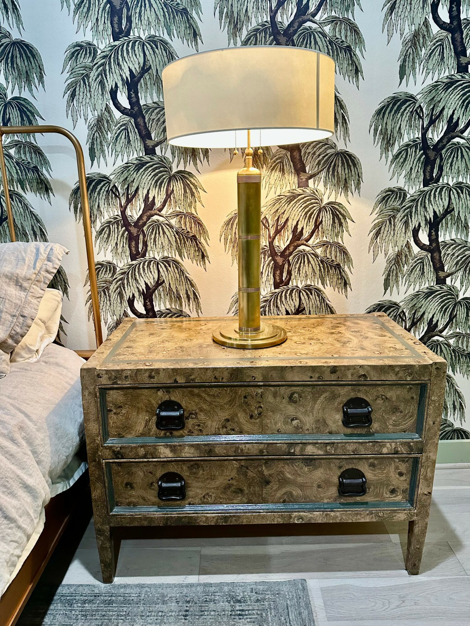 Wooden bedside table with a lamp on top, against a tropical leaf patterned wallpaper, next to a bed with pillows.