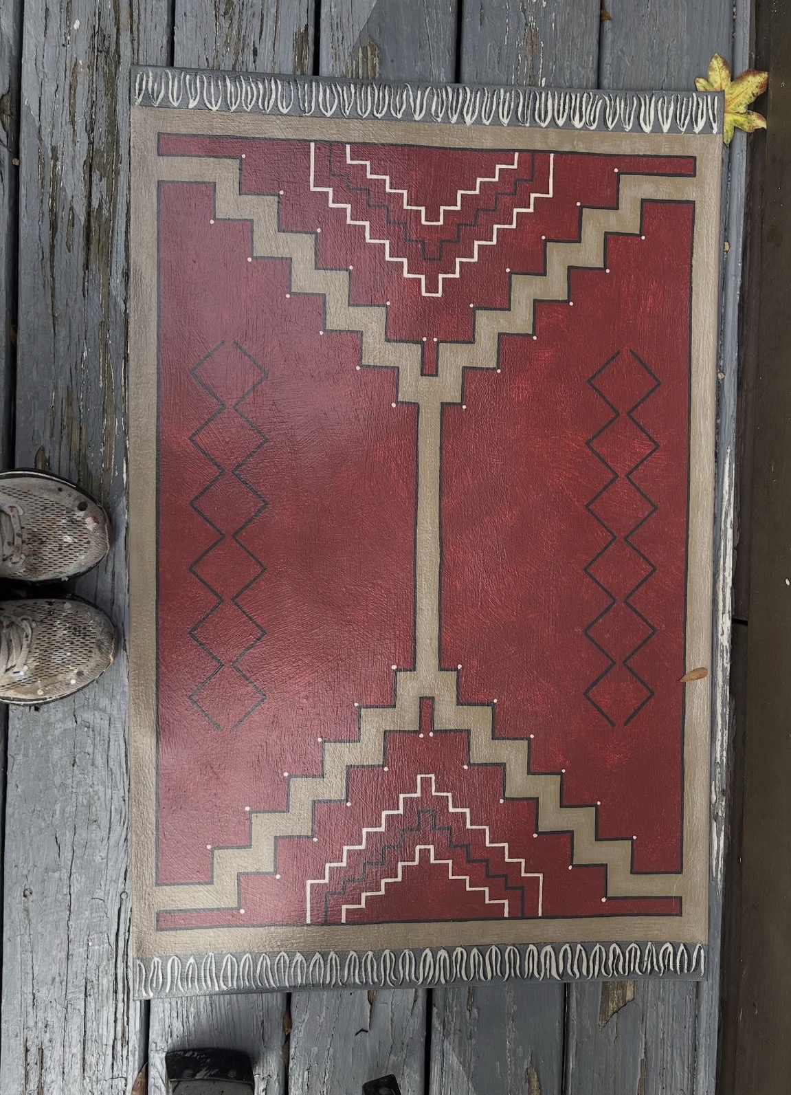 A geometric patterned rug with red, beige, and brown colors, placed on a wooden floor, with a yellow flower in the top right corner.