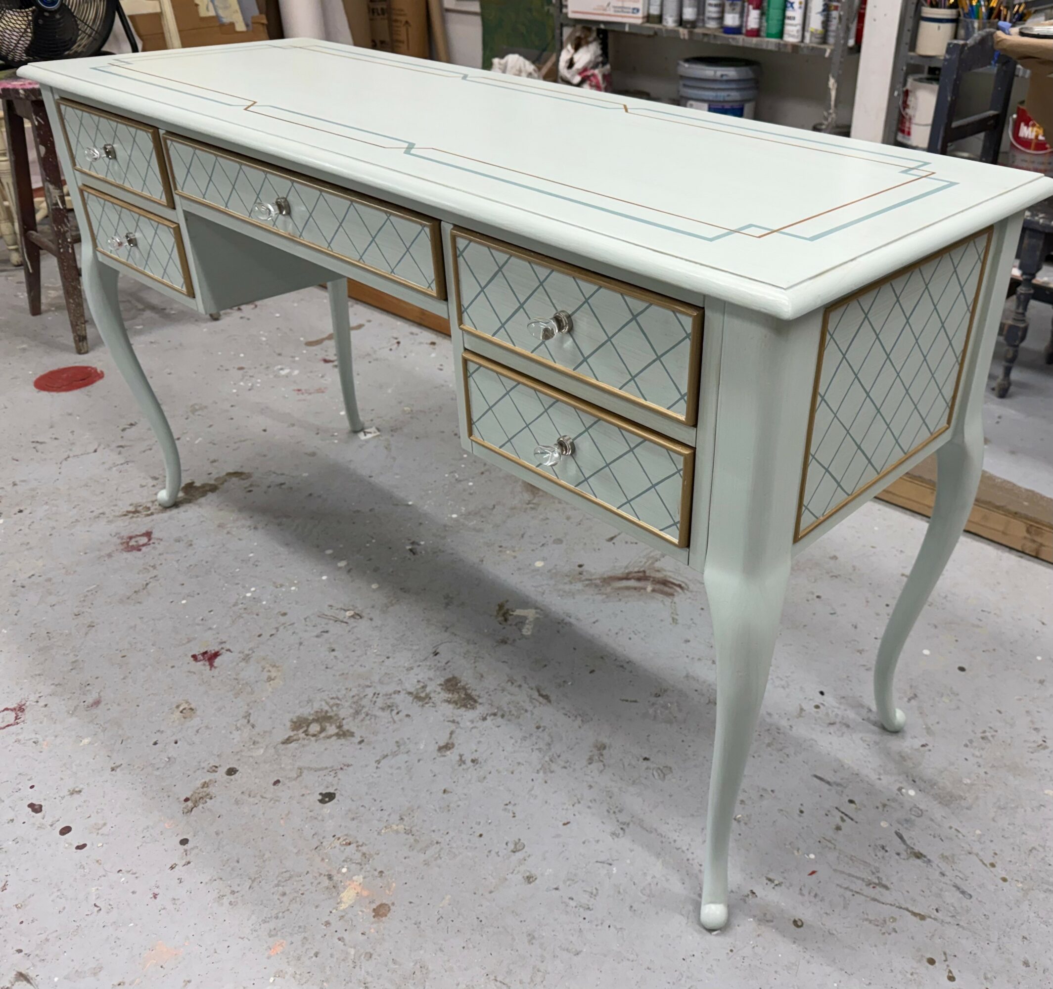 A vintage-style light green desk with gold accents and diamond-patterned drawer fronts, standing on curved legs in a workshop.