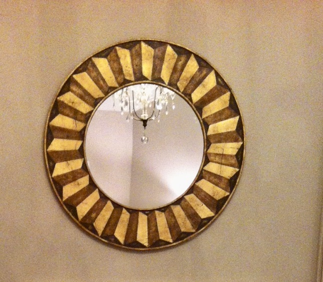 Decorative round mirror with a gold geometric frame hanging on a wall, reflecting a chandelier and ceiling light.
