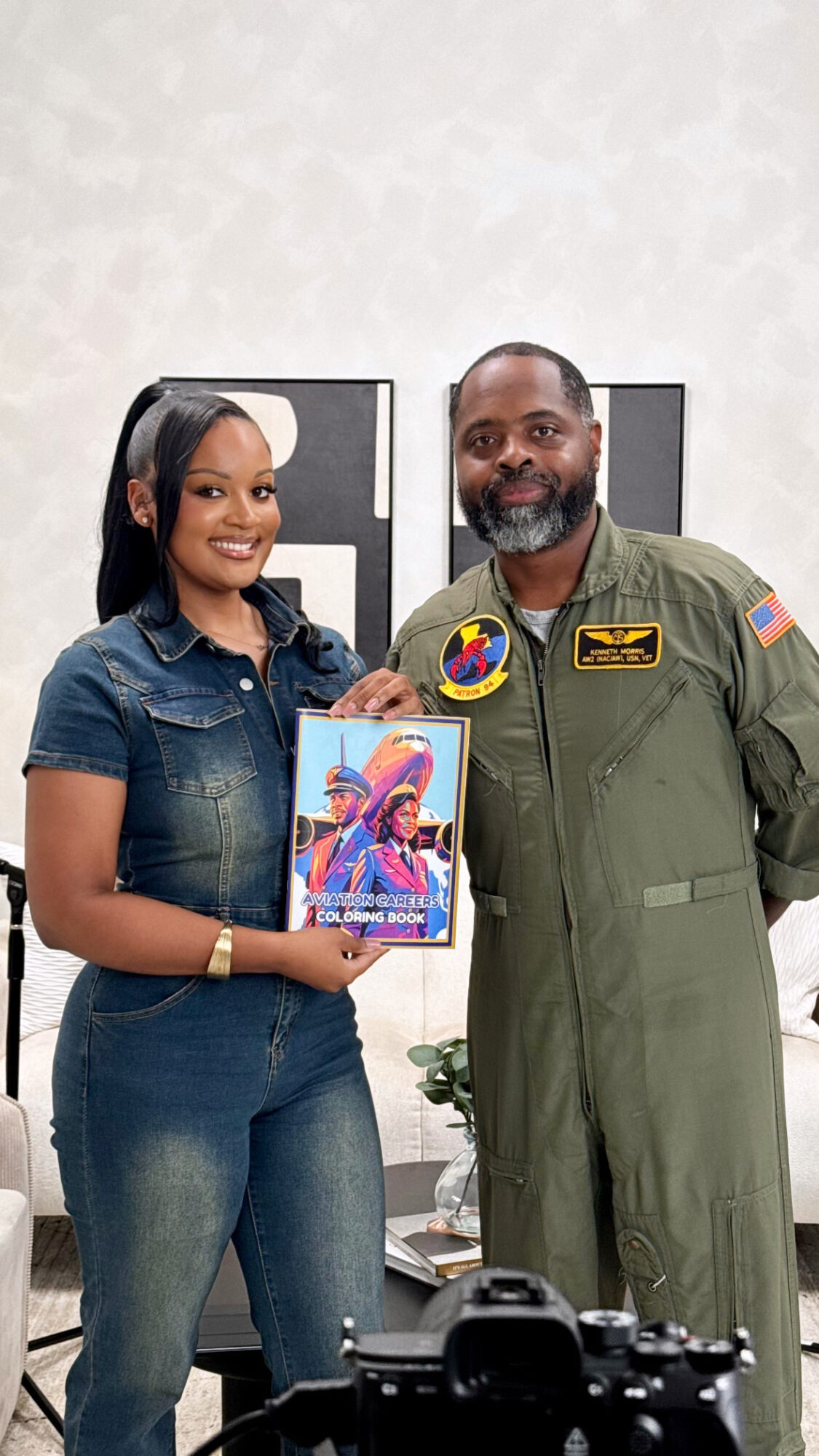 A woman and a man stand together indoors, holding a colorful book, smiling at the camera.