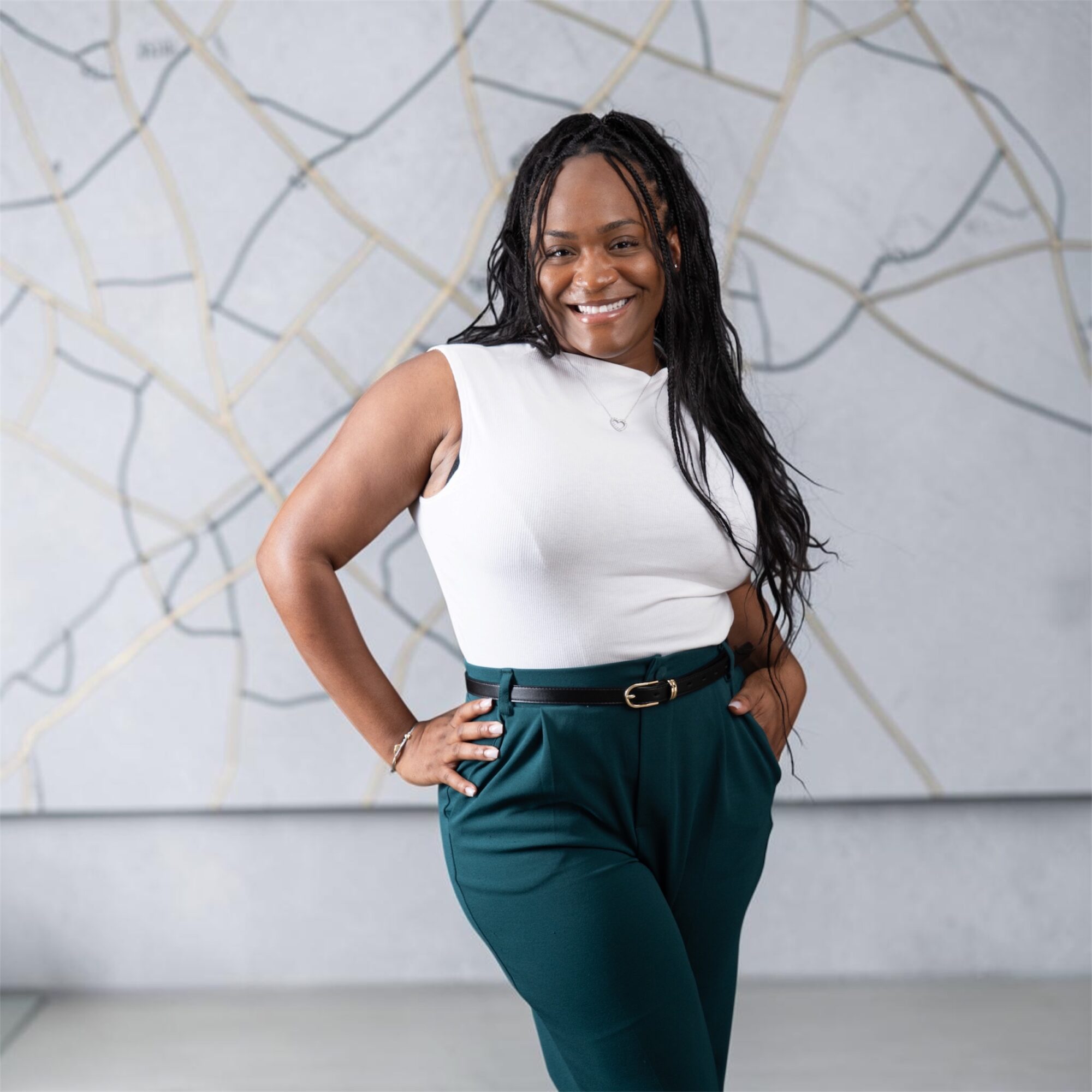 Smiling woman with long dark hair in a white sleeveless top and teal pants, standing in front of an abstract wall.