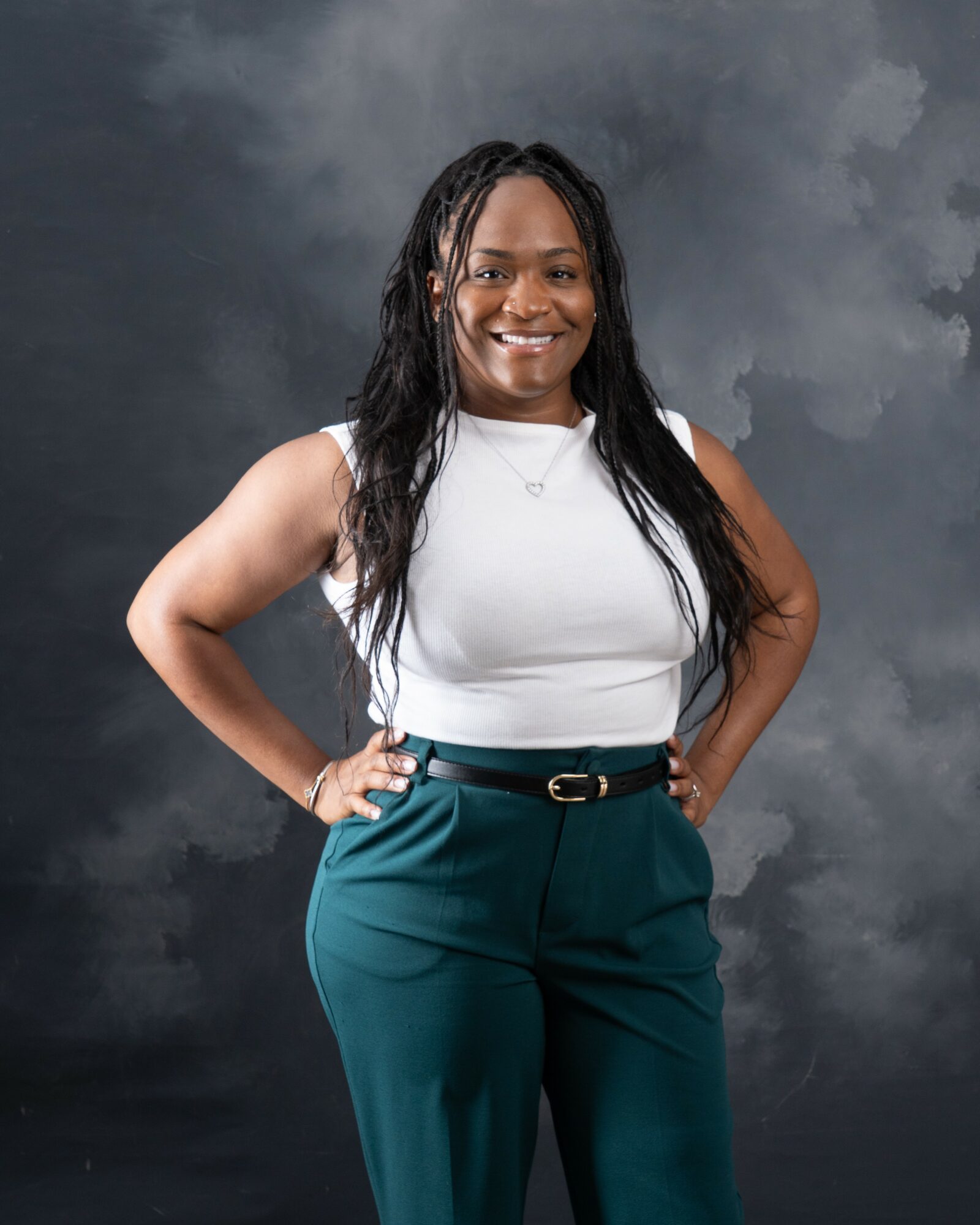 Woman with long dark hair smiling, wearing a white sleeveless top and teal pants, standing with hands on hips against a cloudy background.