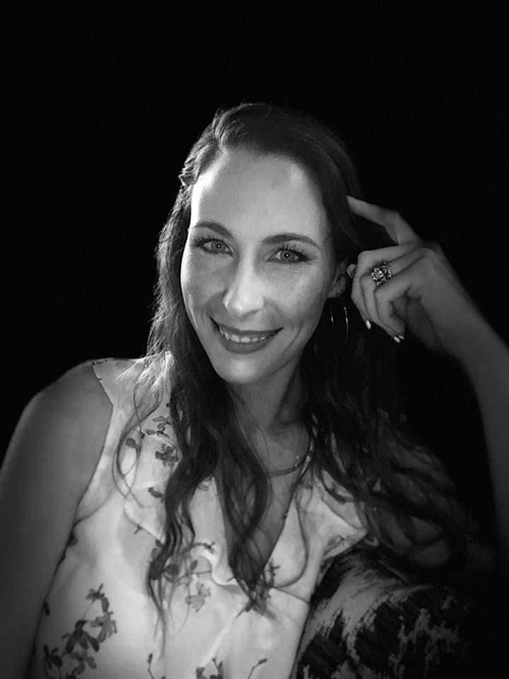 Smiling woman with long hair touching her temple, wearing a floral top, against a dark background.