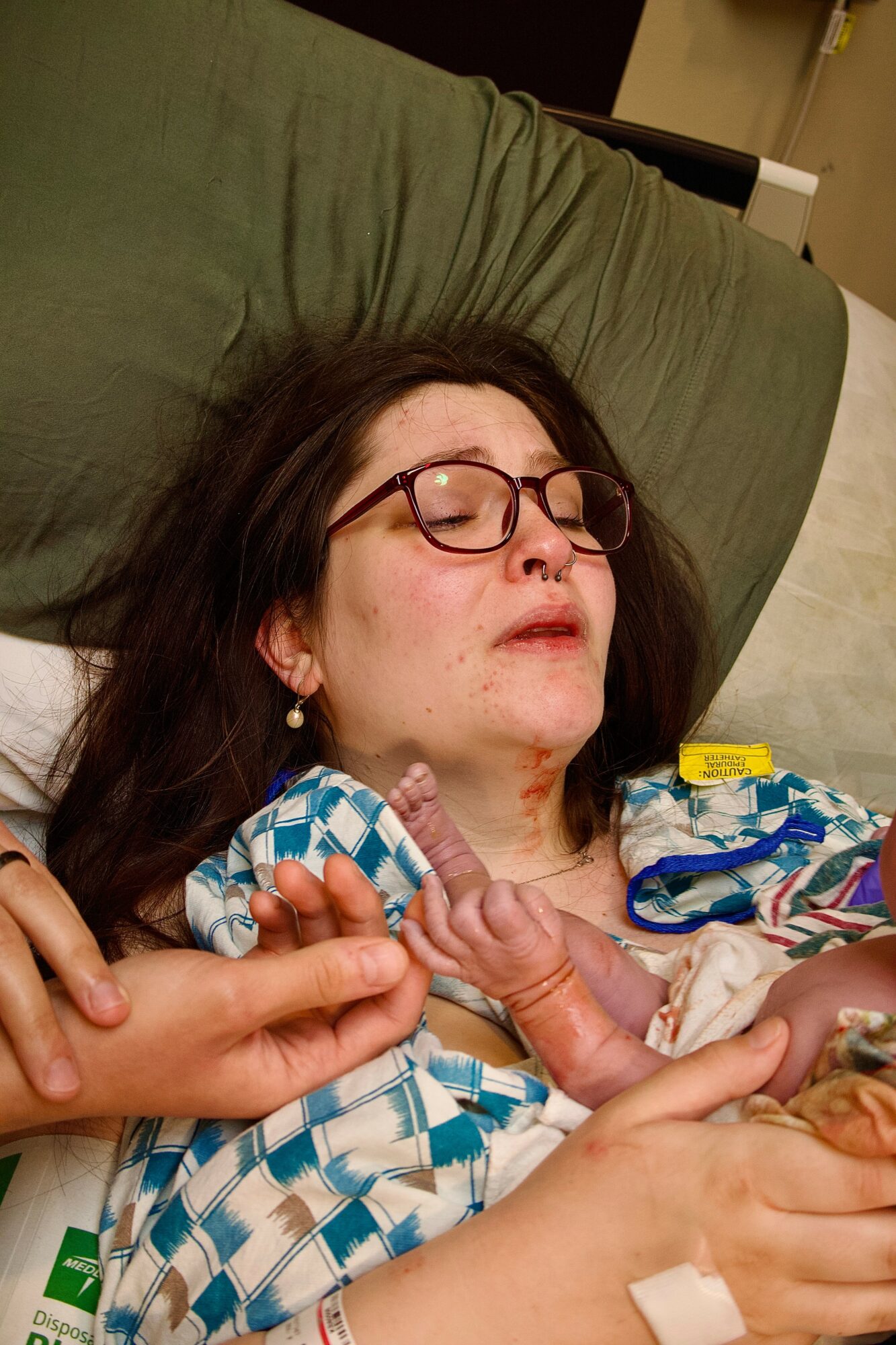 Woman with glasses lying in hospital bed, connected to medical devices, with a hand holding her finger.