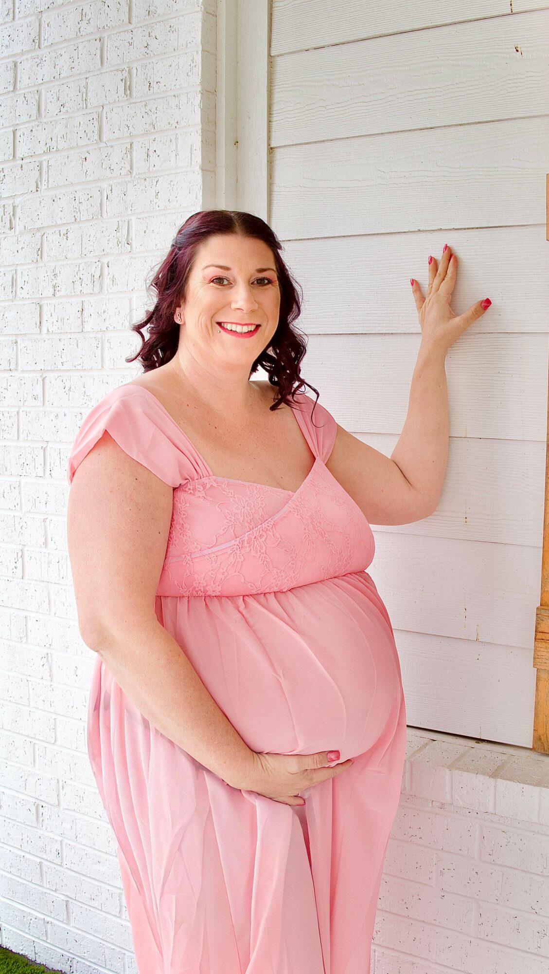 Pregnant woman in pink dress smiling, standing against white wall, hand on wall, holding belly.