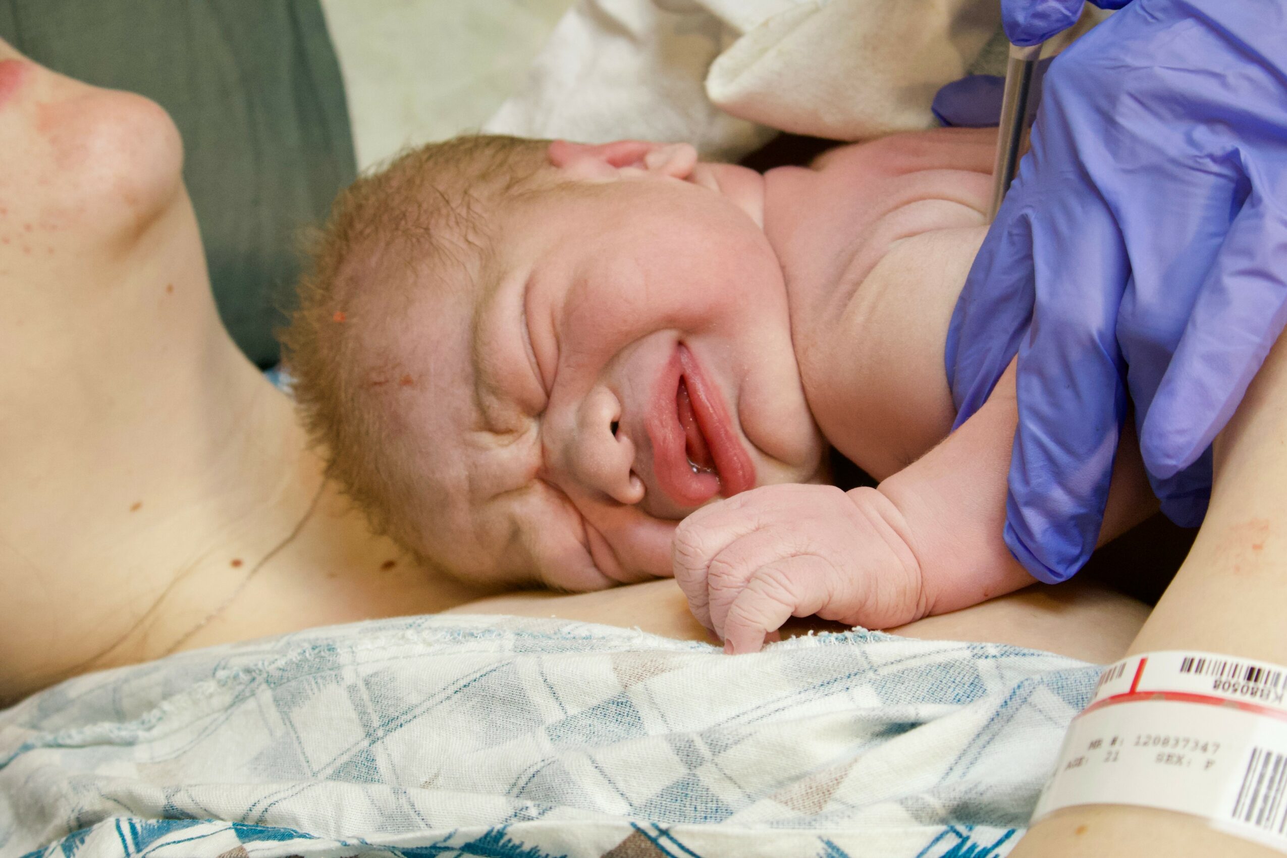 Newborn baby crying, lying on a hospital bed, with medical gloves and equipment nearby.
