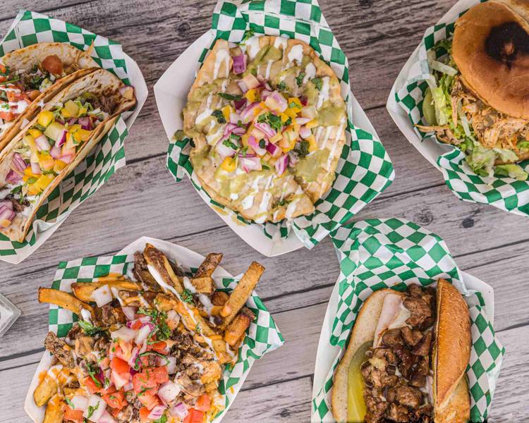 Five different tacos and a burger on a wooden table, served in green and white checkered paper baskets.
