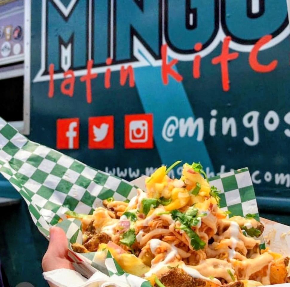 Close-up of a tray of loaded tacos with toppings, in front of a food truck with social media icons and text.