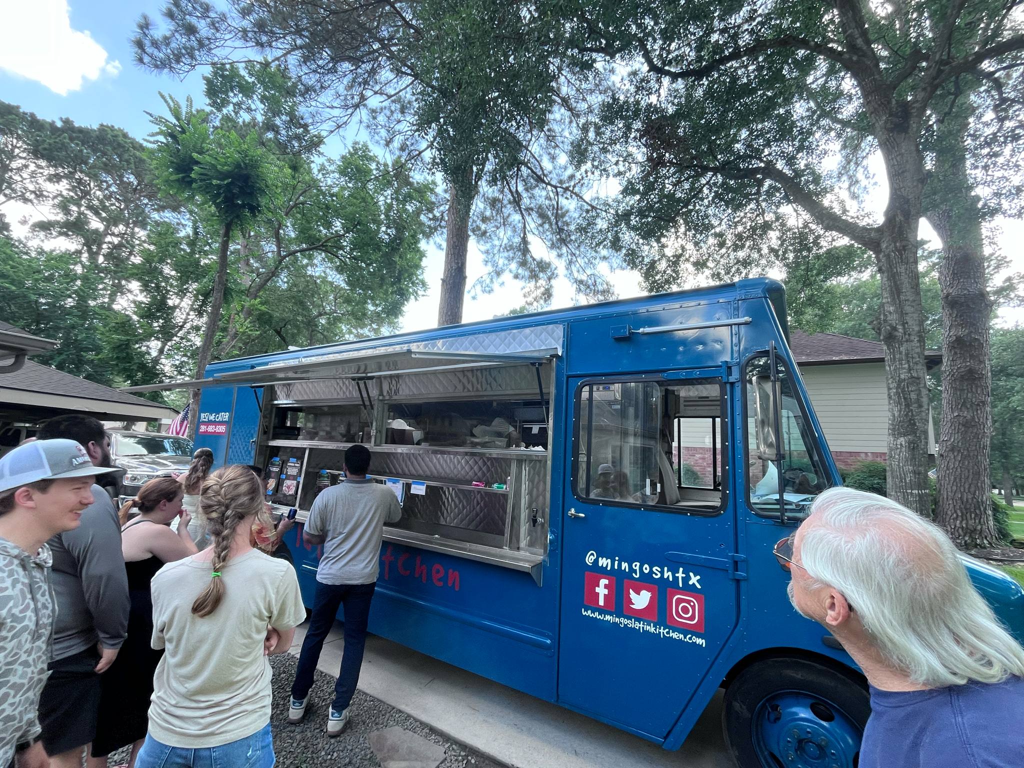 People line up at a blue food truck outdoors with trees in the background, some wearing hats and masks.