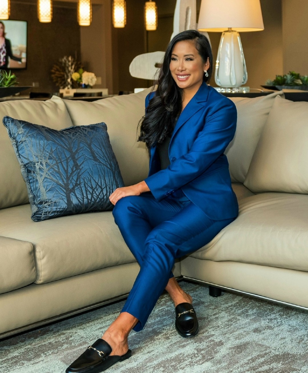 Woman in blue suit sitting on a beige sofa, smiling, with a large blue pillow, in a cozy living room.