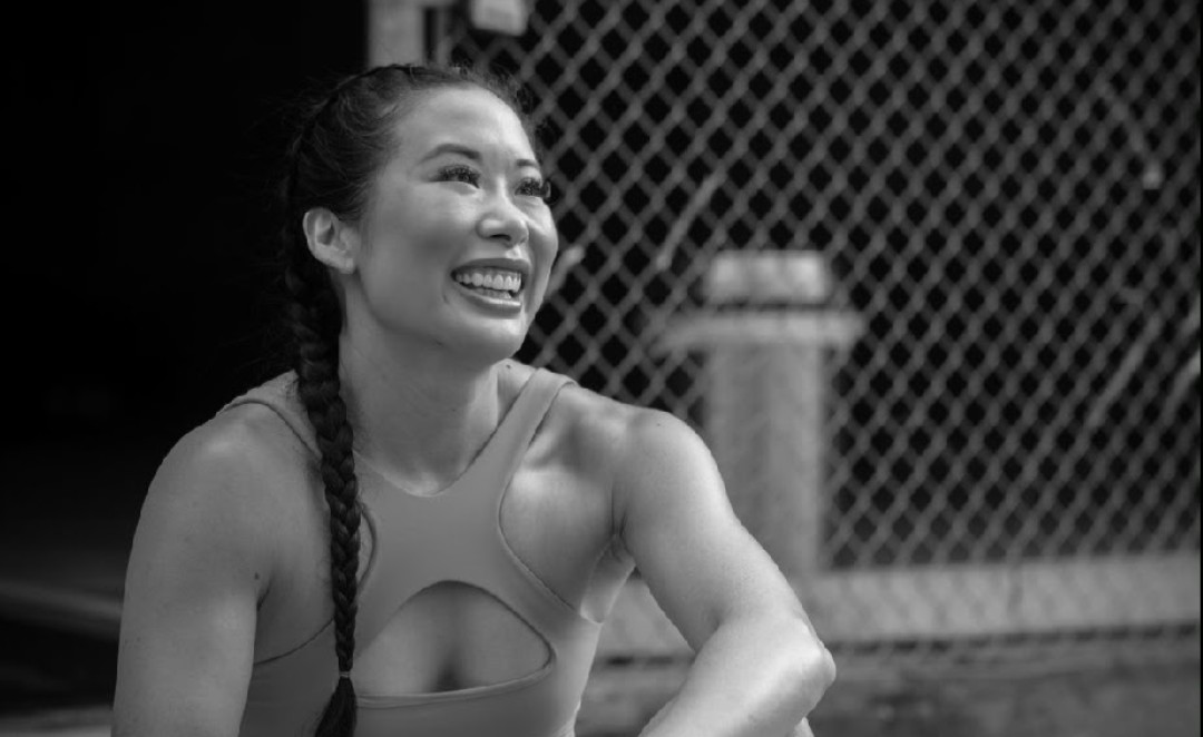 Smiling woman with braided hair outdoors near a chain-link fence, wearing a sports top.