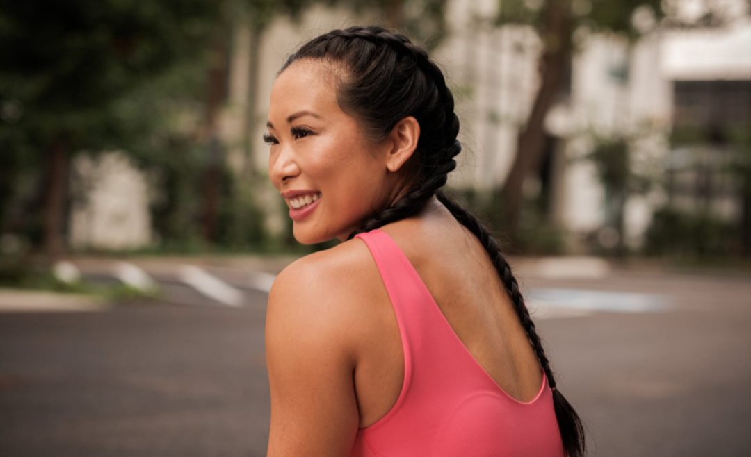 Smiling woman with braided hair in pink athletic top outdoors, background of trees and buildings