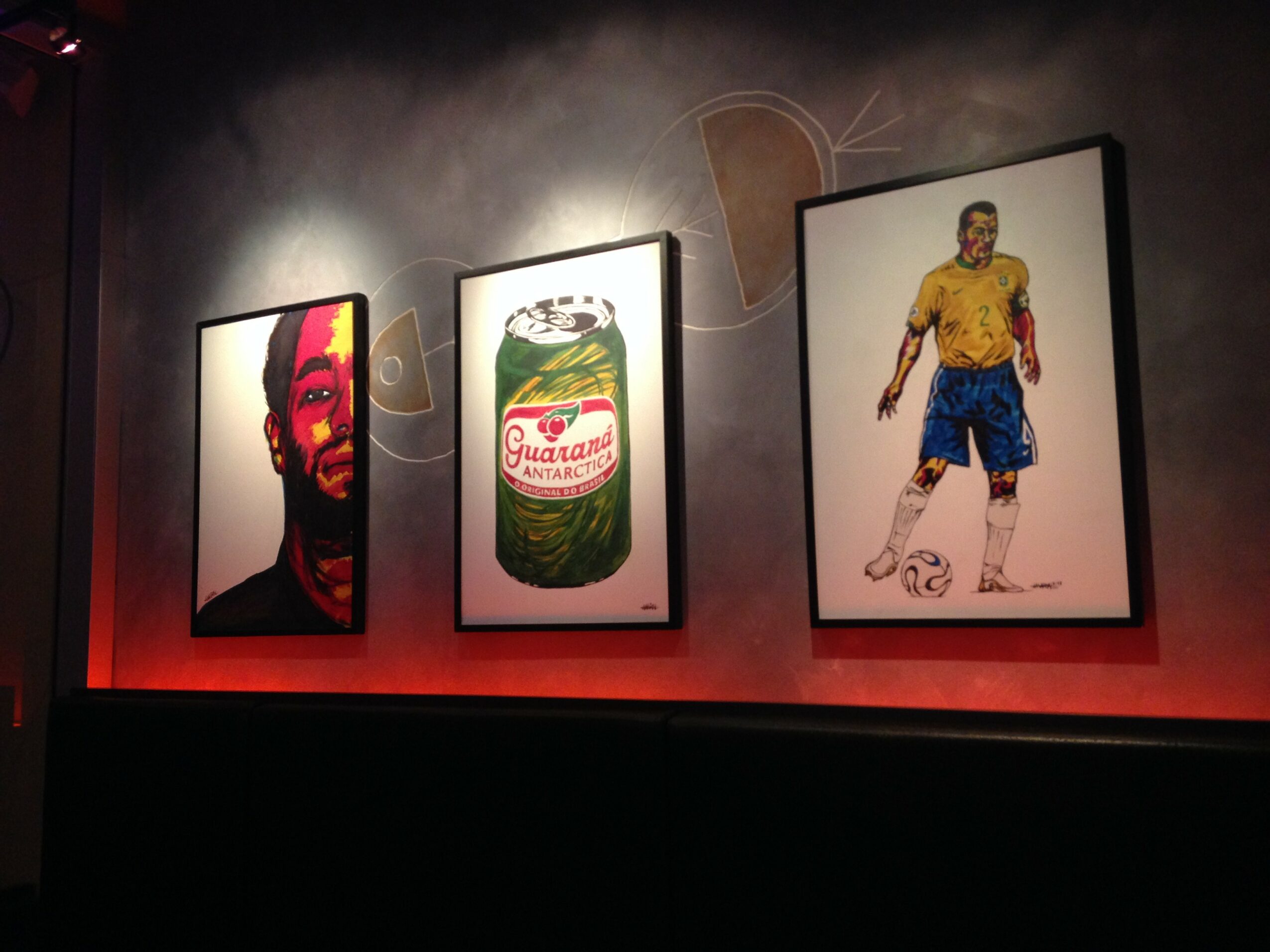 Three framed images on a wall: a person with a red face, a can of Guaraná soda, and a soccer player in yellow and blue uniform.