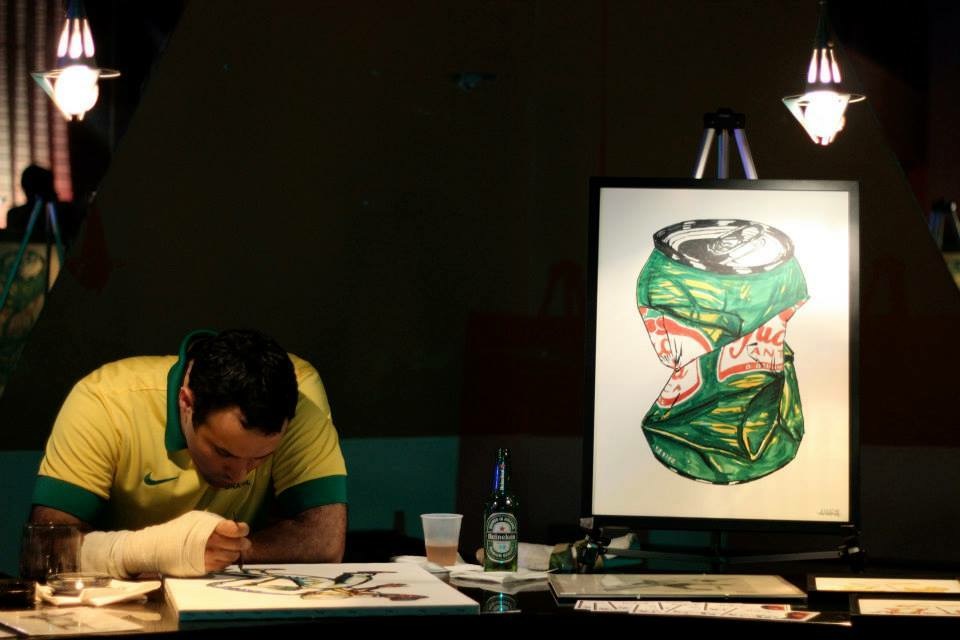 Person in yellow shirt leaning over table with artwork, illuminated display with abstract green and red drawing, bottles and books nearby.