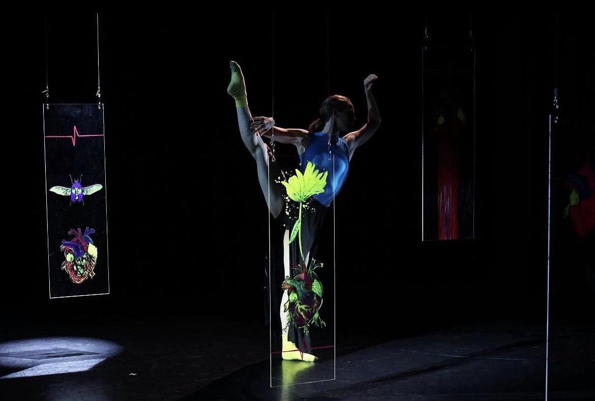 Performer in a blue costume poses with one leg raised, surrounded by vertical panels with colorful designs, on a dark stage.