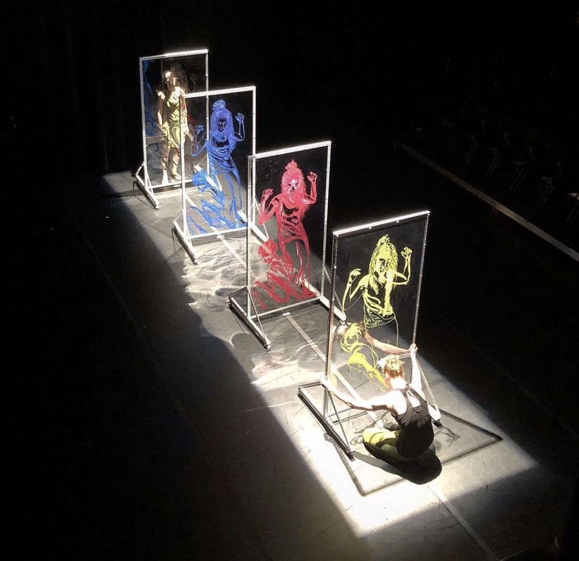 Person sitting on the floor in front of colorful illuminated art panels with figures, in a dark room.