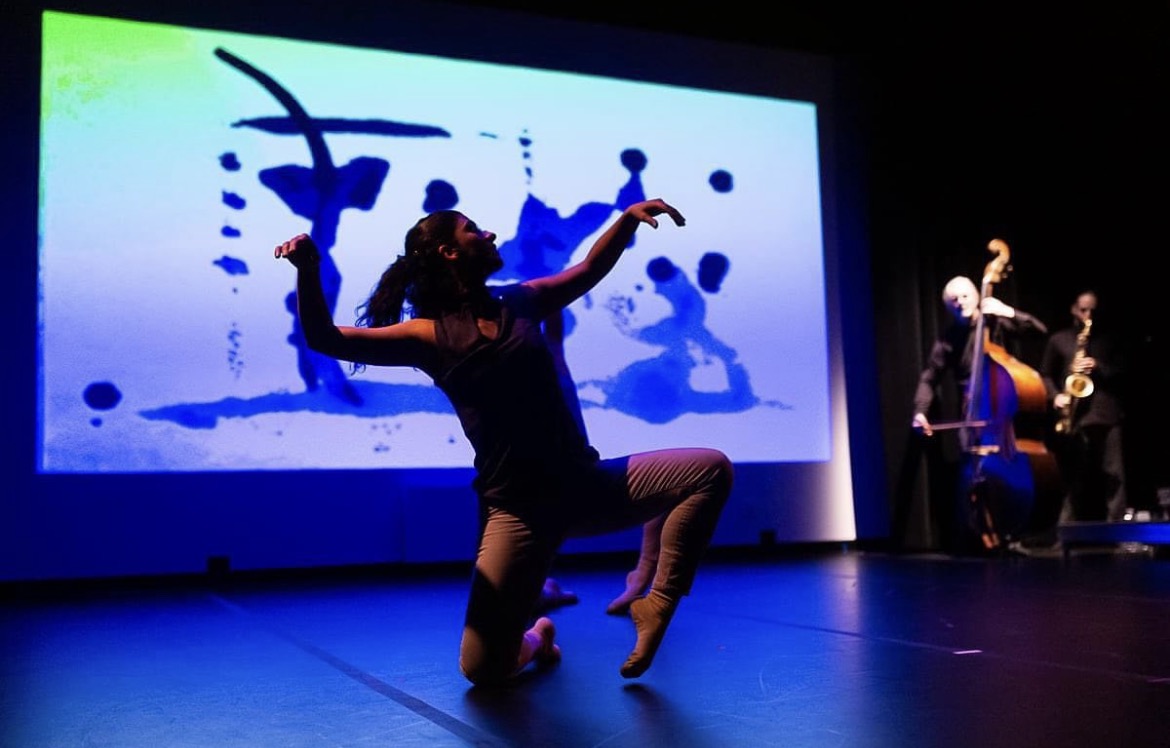 Performer dancing on stage with a large screen displaying abstract blue and black ink splashes behind them.