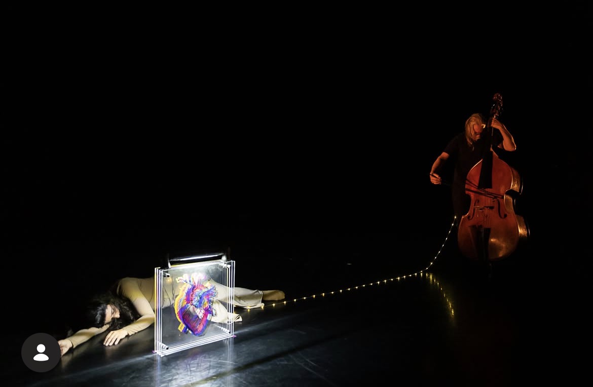 Person lying on the floor with a box and a colorful object, and a person playing a double bass on a dark stage.