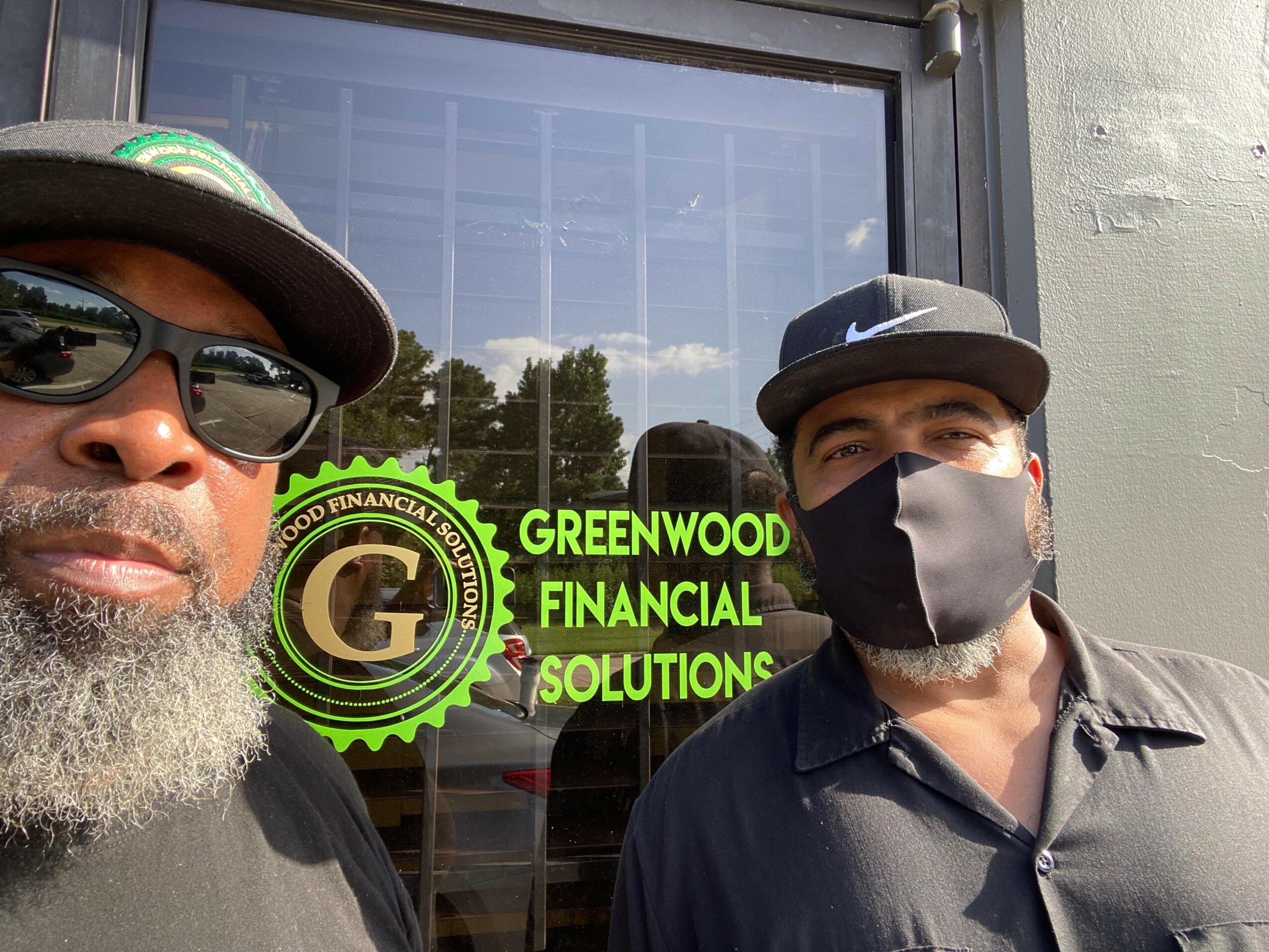 Two men wearing hats and face masks stand outside a business with a sign reading 'Greenwood Financial Solutions.'