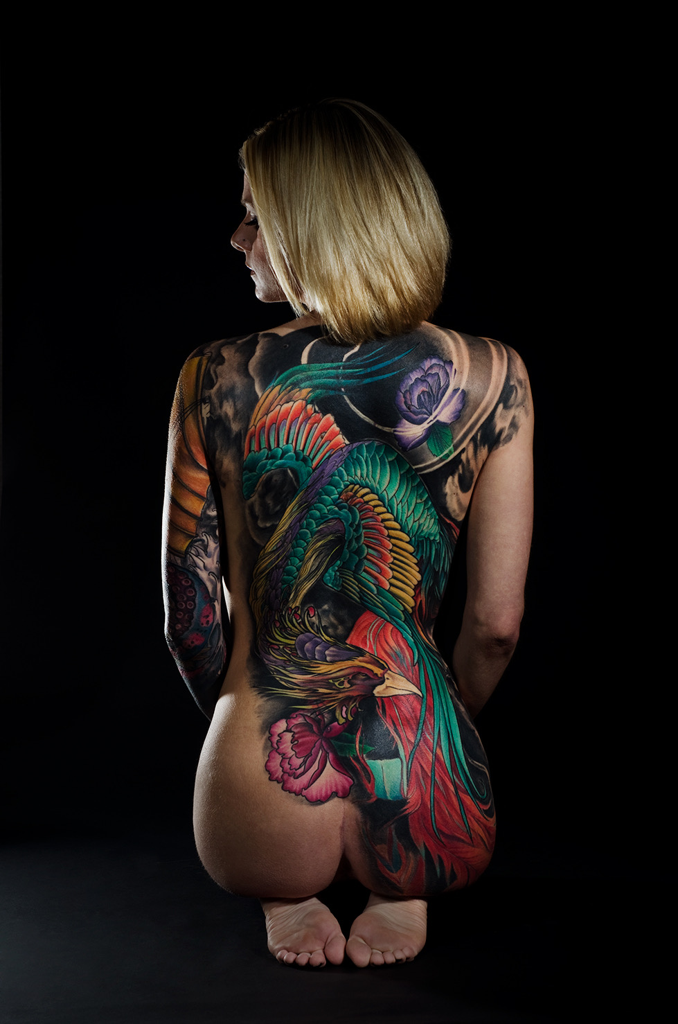 Person with colorful tattoos on back and arms, kneeling with back facing camera, head turned to side, black background.