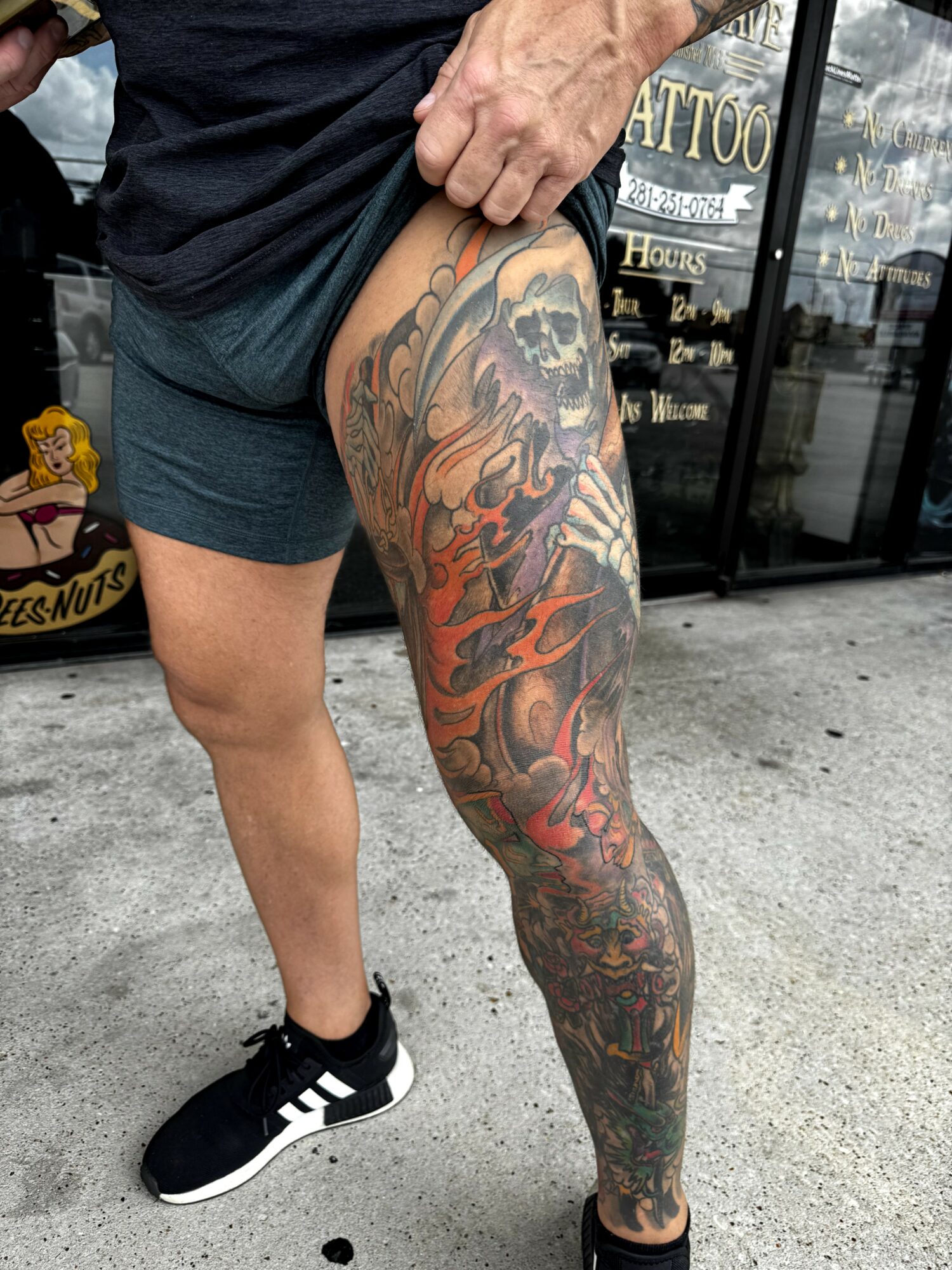 Person with a large, colorful tattoo covering their right leg, standing outside a tattoo shop, wearing black shorts and sneakers.