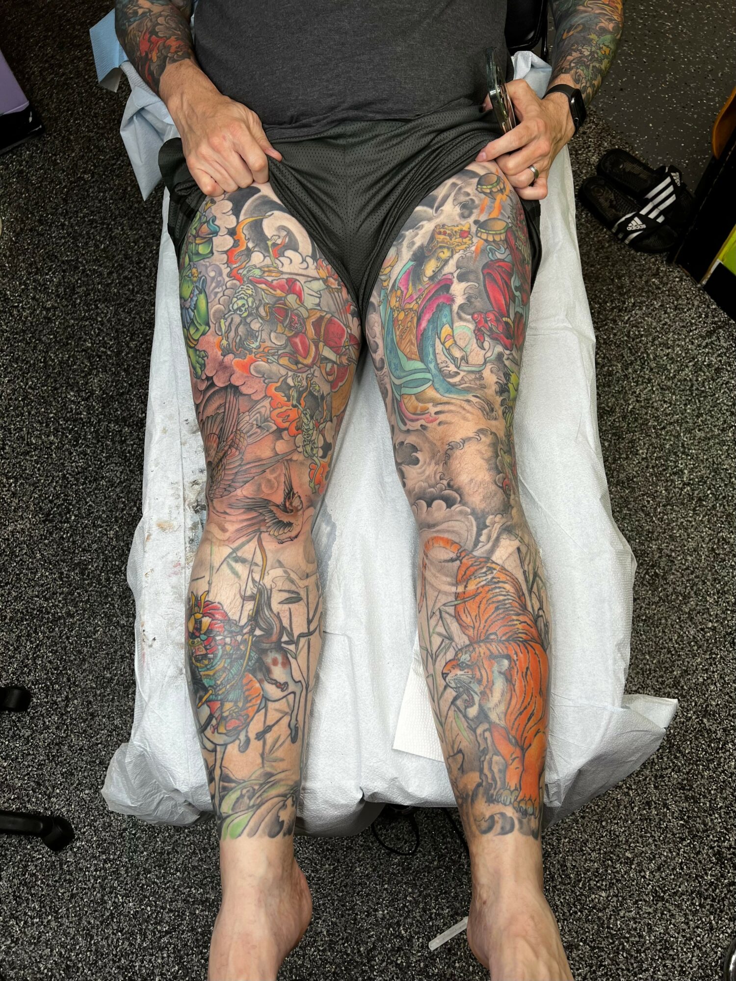 Person with colorful tattoos on legs sitting on a chair, hands resting on thighs, in a room with black carpet.