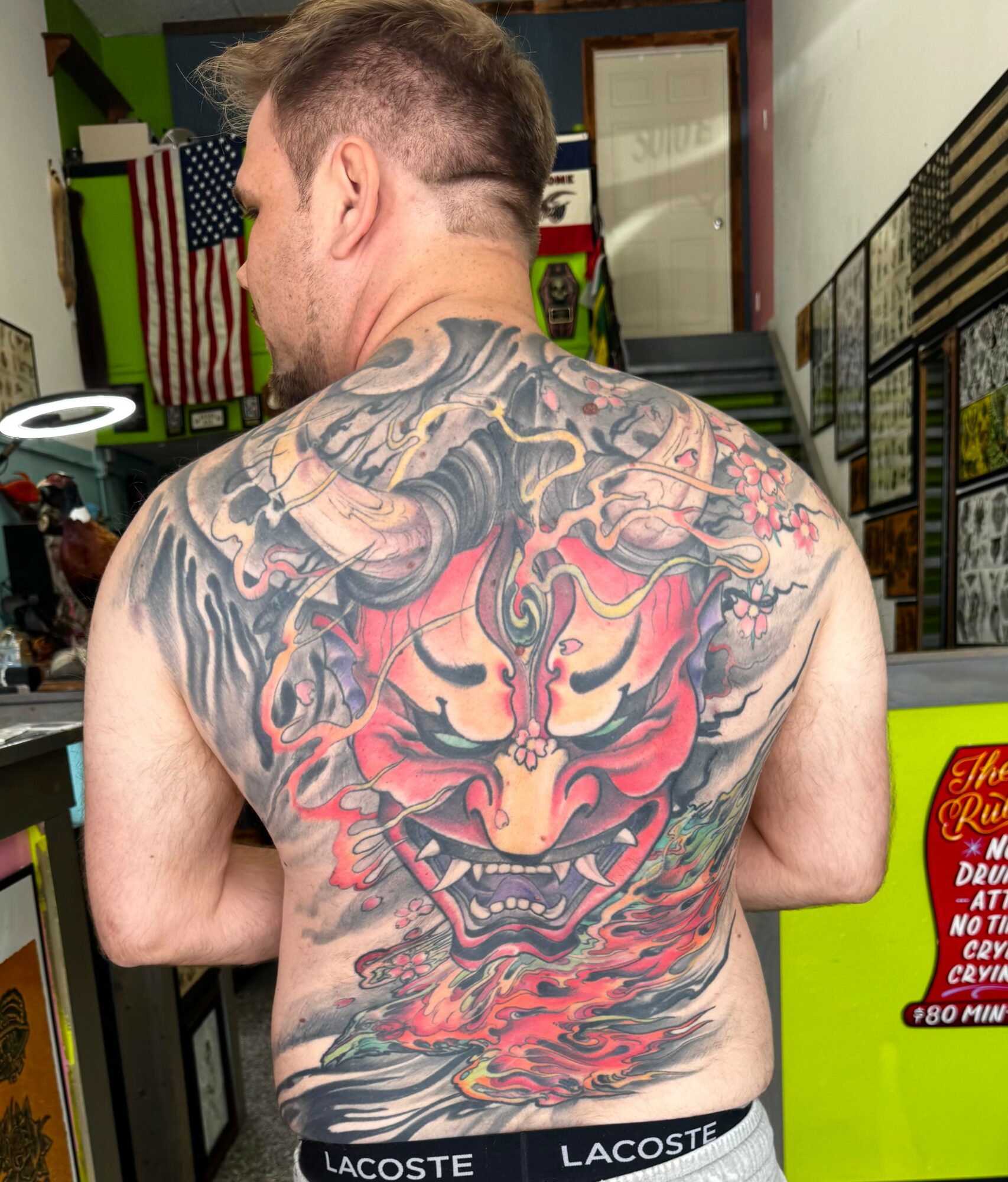 Back view of a man with a large, colorful tattoo of a demon face on his back, in a store setting.