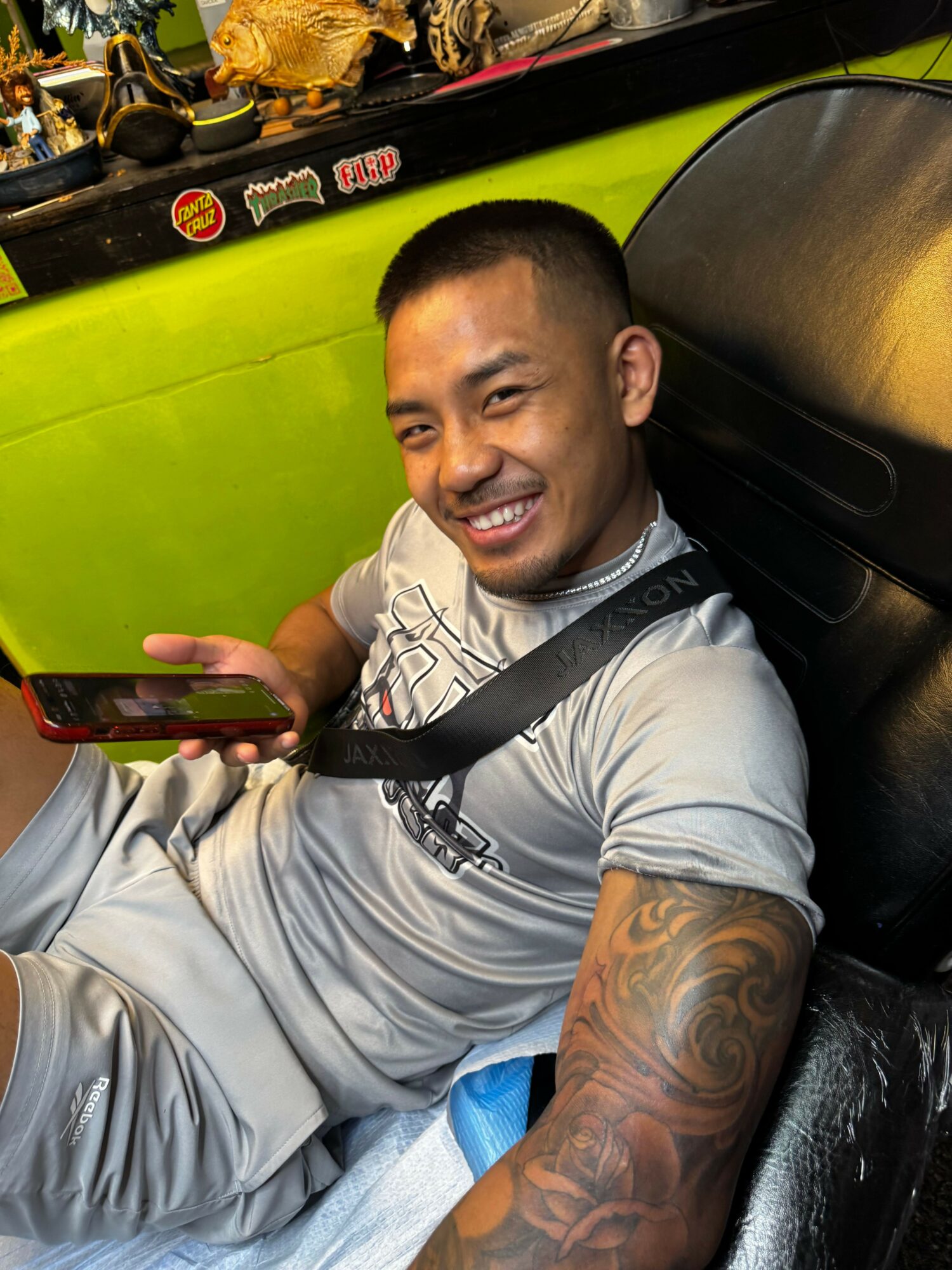 Man with tattoos on arm, smiling, holding a phone, sitting on a black leather seat, with a green background.
