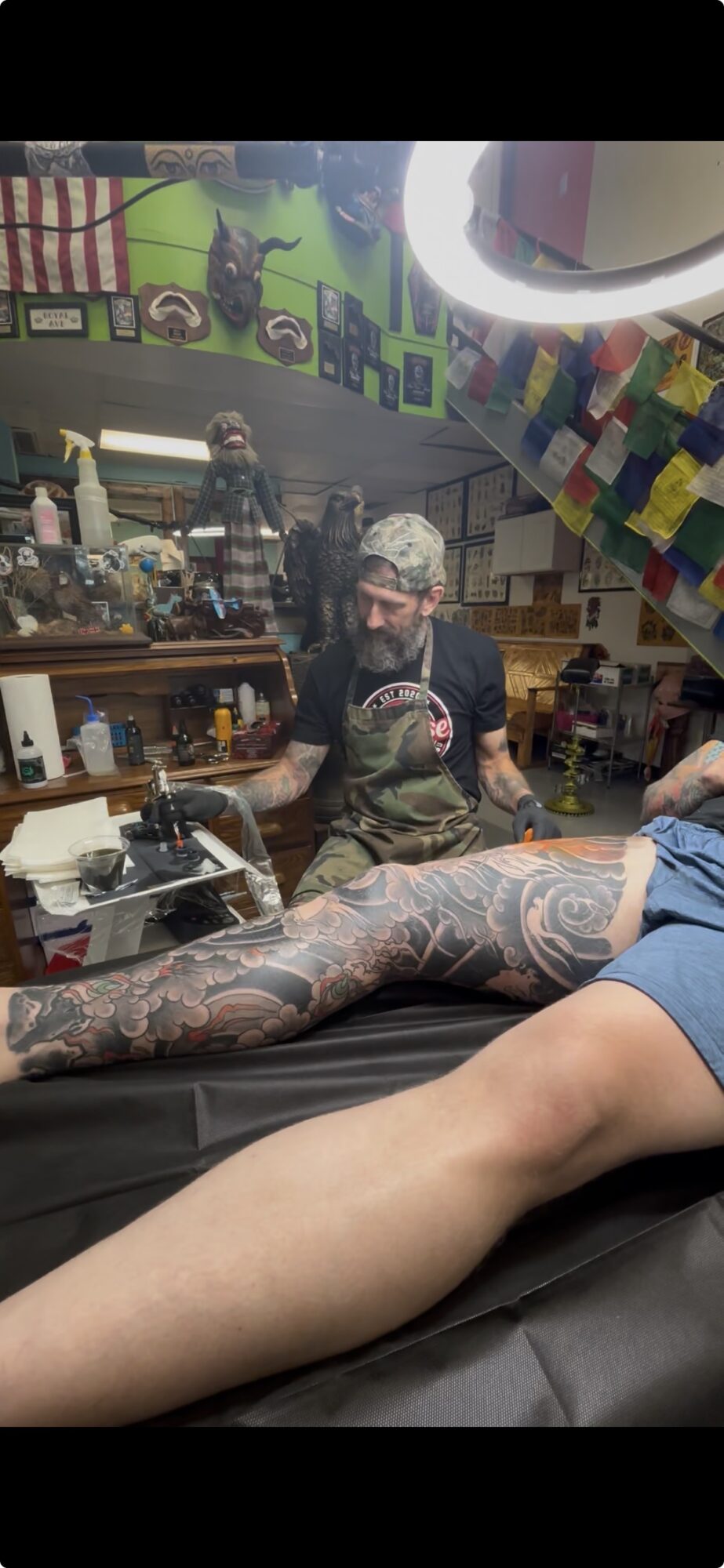 Person with tattooed arm lying on table, tattoo artist working in a colorful shop with artwork on walls.
