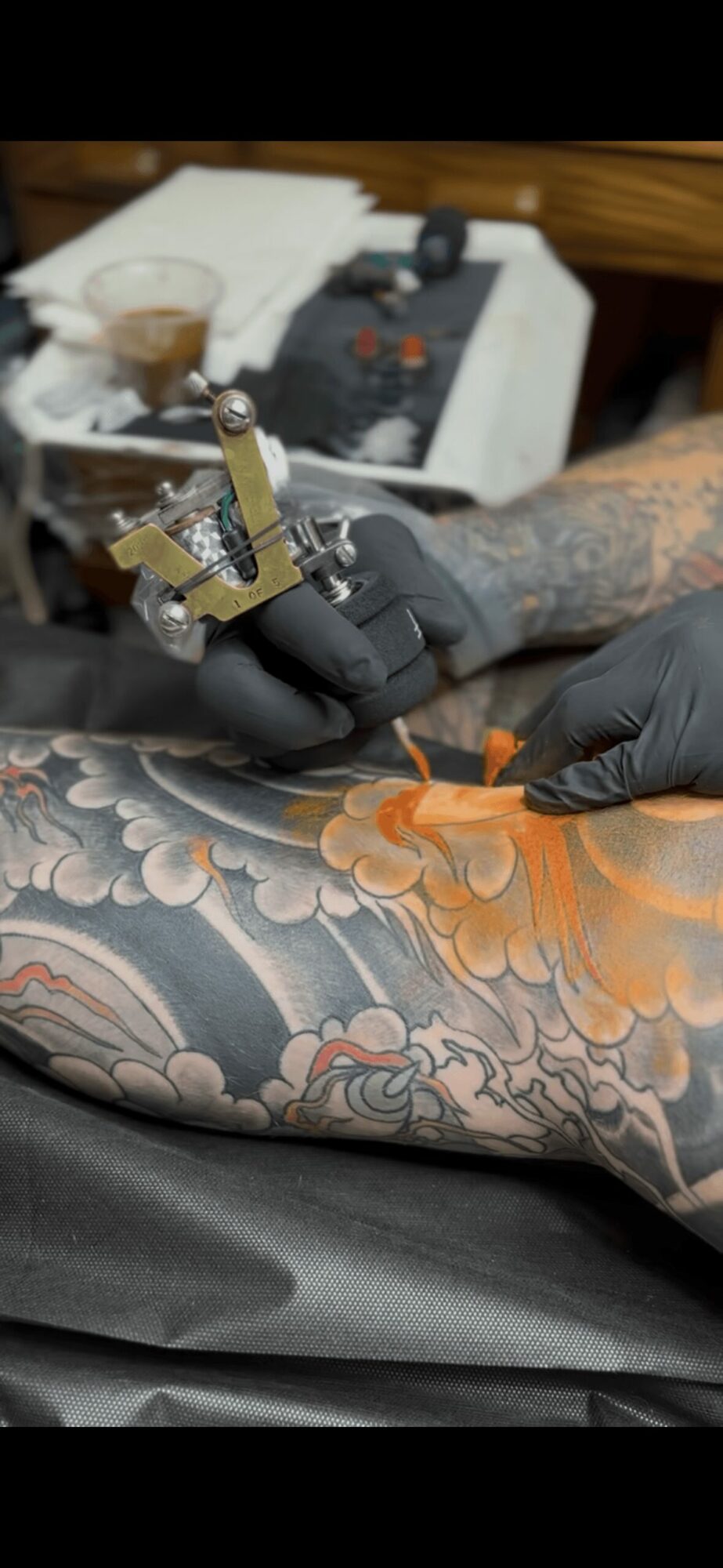 Tattoo artist's hand with black glove tattooing a colorful floral design on a person's arm, tattoo machine in hand.