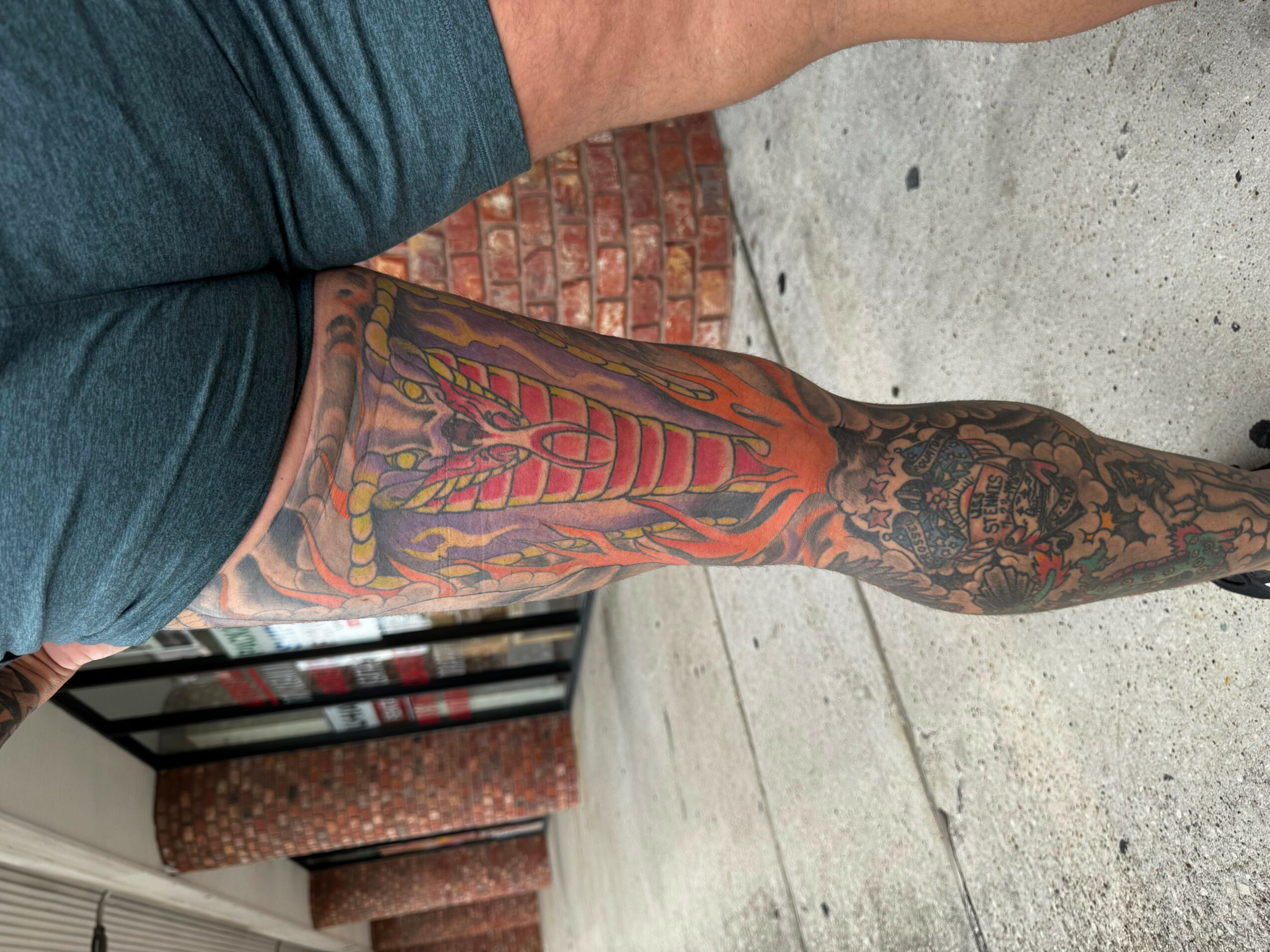 Person with a colorful tattoo sleeve on their arm, standing on a concrete sidewalk near a brick wall.