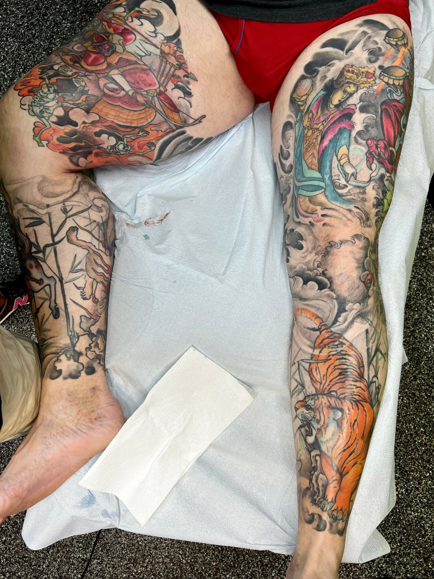Person lying on a surface with colorful tattoos on both arms and legs, wearing red shorts and a black shirt, with a white cloth nearby.