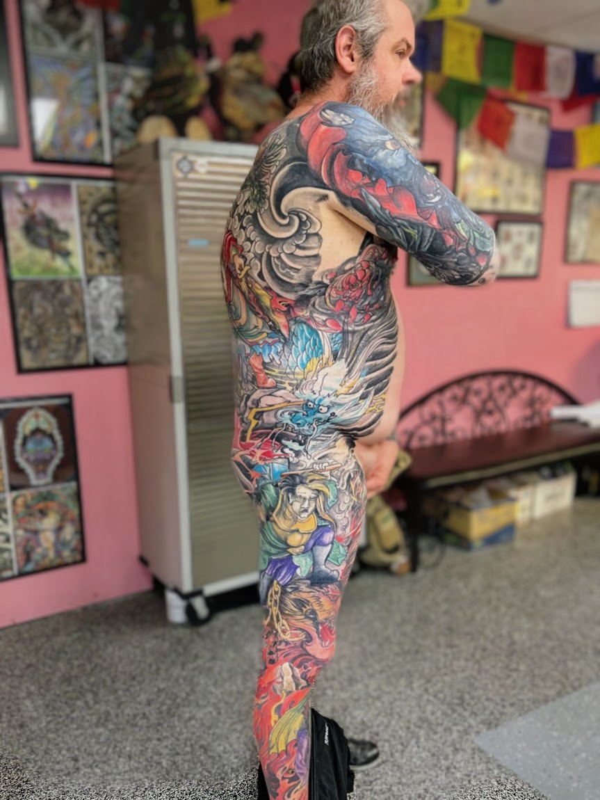 Side view of a tattooed man with gray hair and beard, standing indoors with pink walls and artwork, arms crossed.