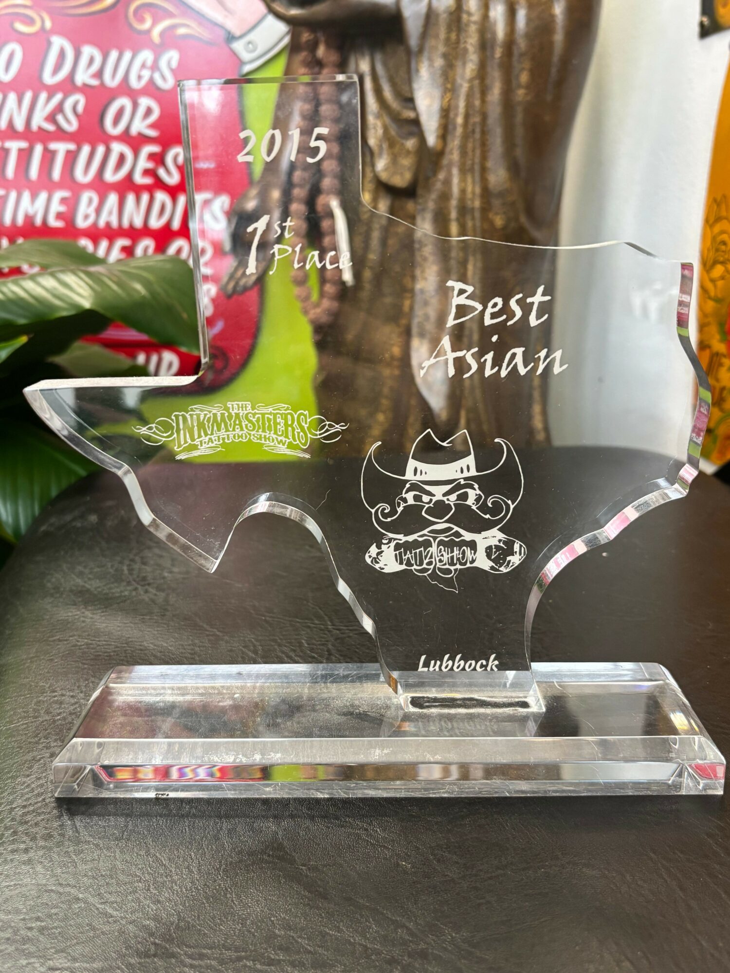 Clear glass award with engraved text and a cartoon character, placed on a black surface with colorful background objects.